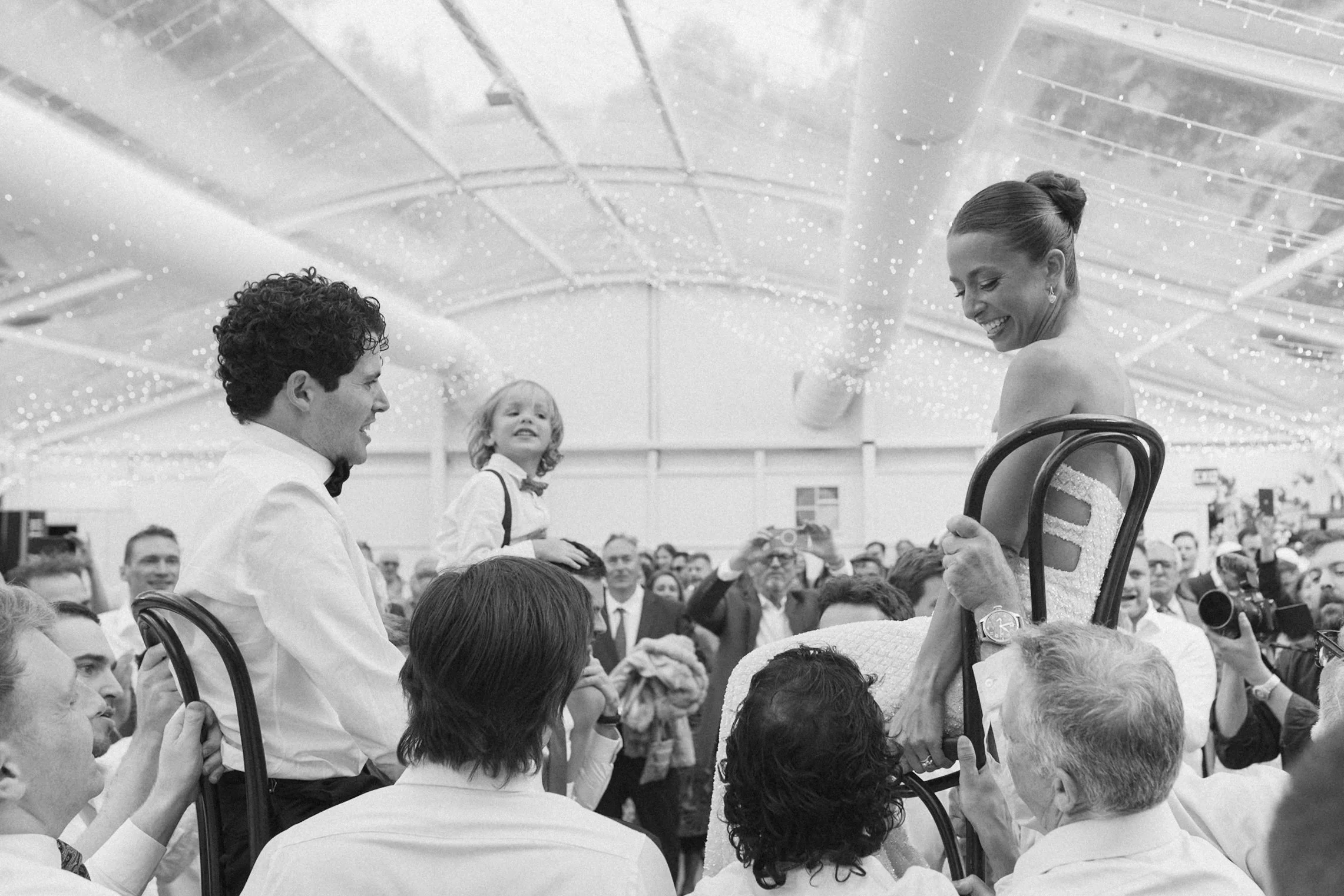 A black-and-white photograph of a woman in a wedding dress and a man in a tuxedo sitting in chairs, surrounded by a crowd of people, with a young girl between them. The woman is smiling and looking at the man while the man looks at her, with the crow