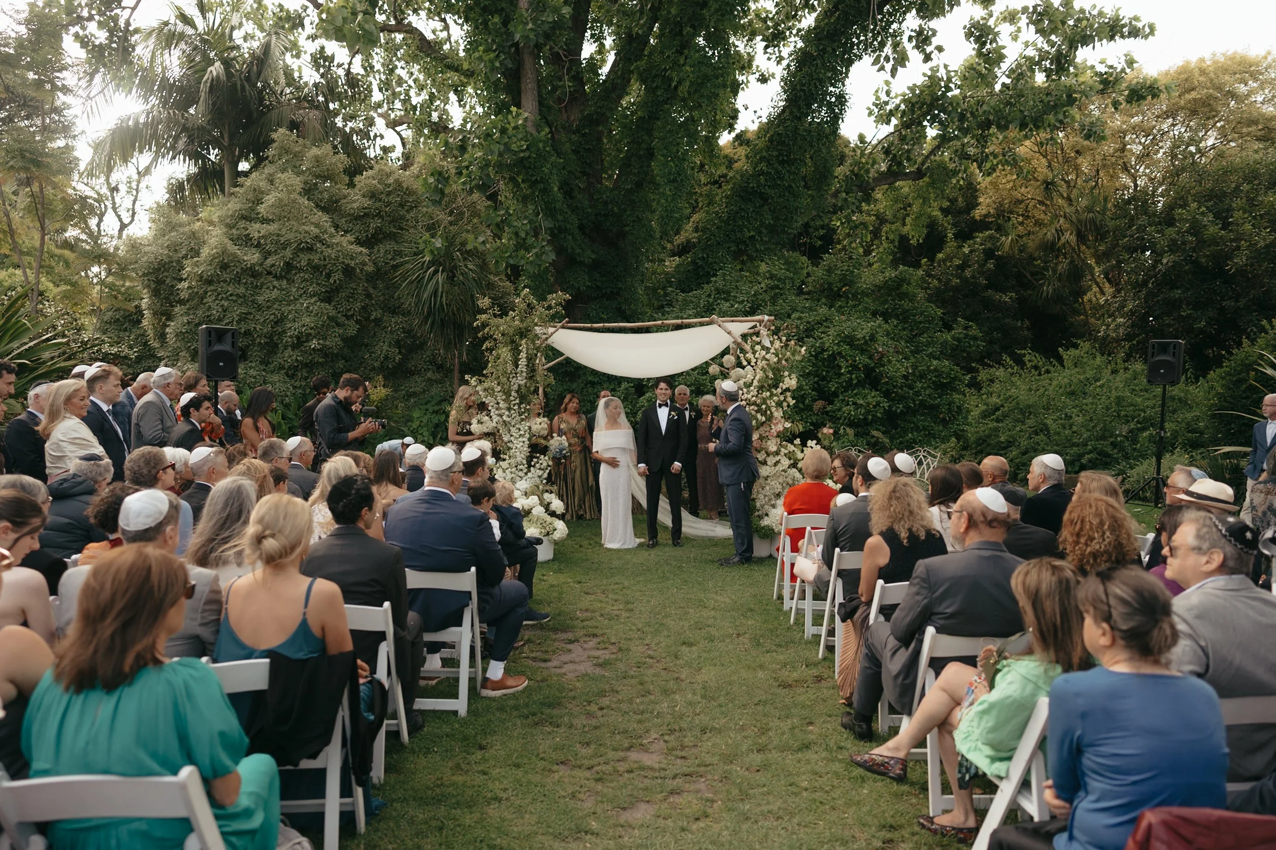 A large outdoor wedding ceremony taking place in a lush green garden with many guests seated and couple standing under a decorated arch.