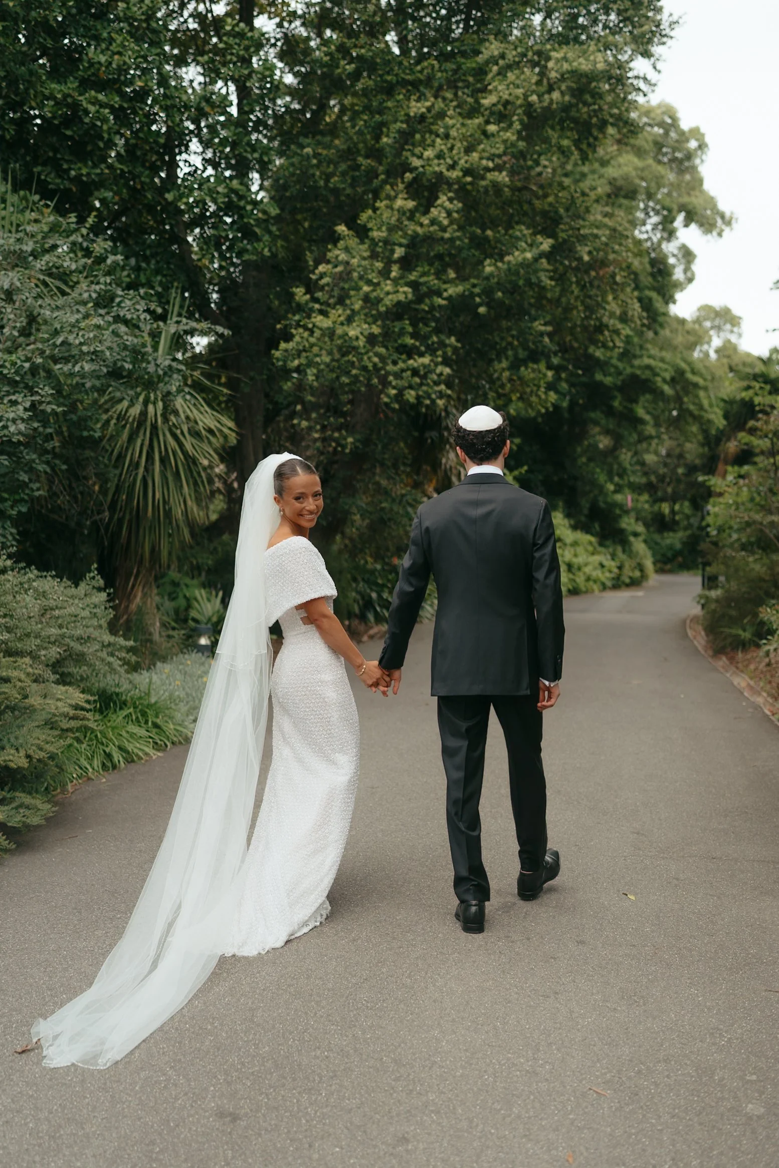 A bride and groom hold hands and walk away on a paved path in a lush green park. The bride is smiling and looking back, wearing a white wedding dress and veil, while the groom is dressed in a black suit and kippah.