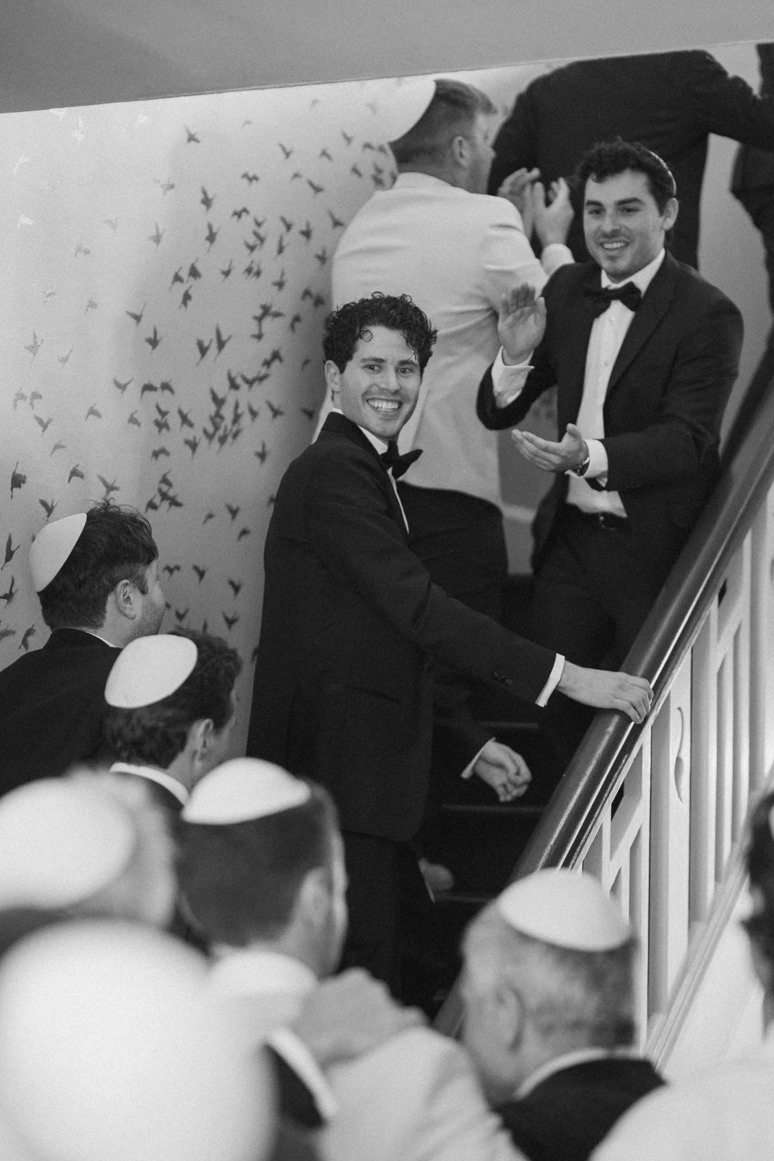 Men dressed in tuxedos and bow ties, some wearing kippahs, ascending a staircase while smiling and waving, with a wall decorated with numerous small bird cutouts in the background.
