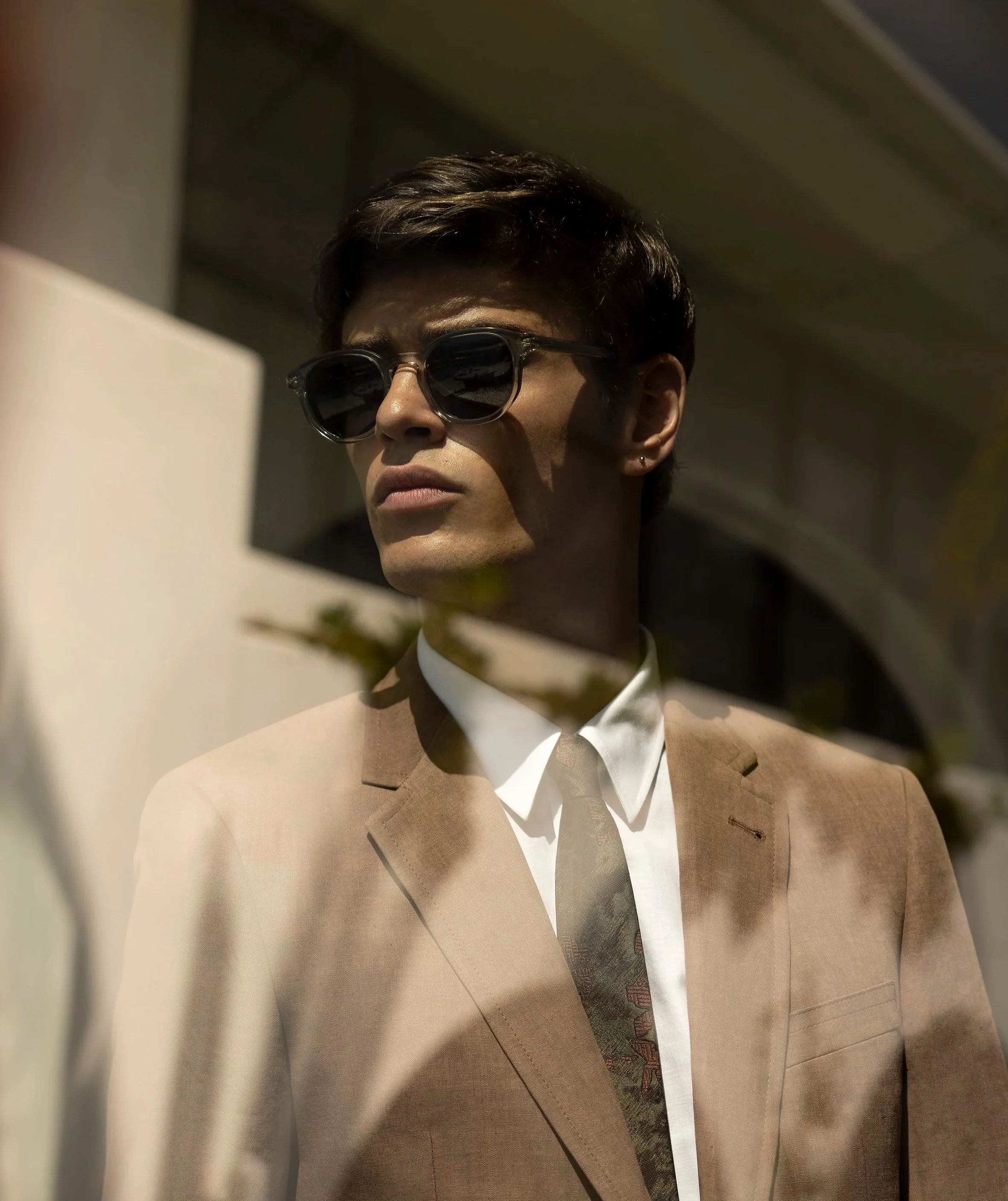 A person with short dark hair wearing sunglasses, a beige suit, and a white shirt, looking sideways at an angle.