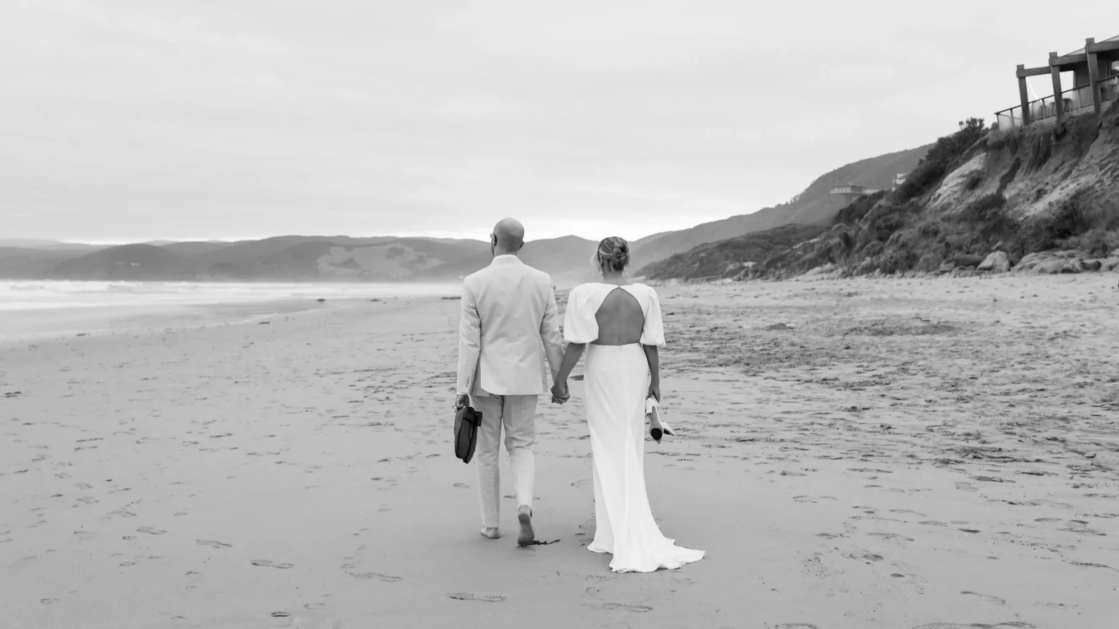 A couple dressed in wedding attire walking hand in hand on a beach, with cliffs and houses in the background.