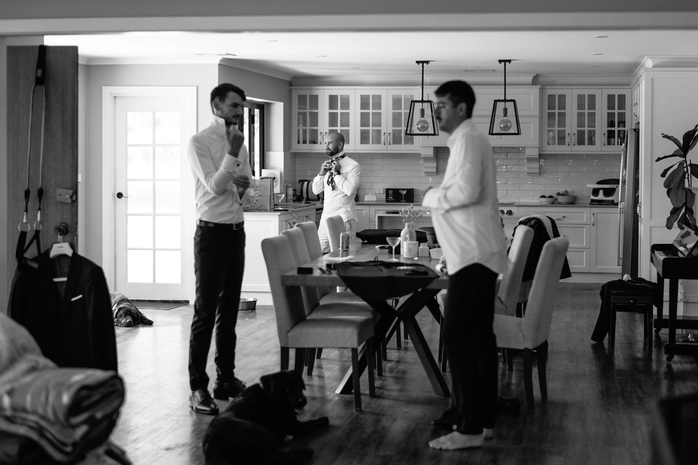 A black-and-white photo of three men in a kitchen. Two men in white shirts are standing and talking, while the third man is in the background, adjusting a bow tie. The kitchen has white cabinets, a countertop with a microwave, and pendant lights hang