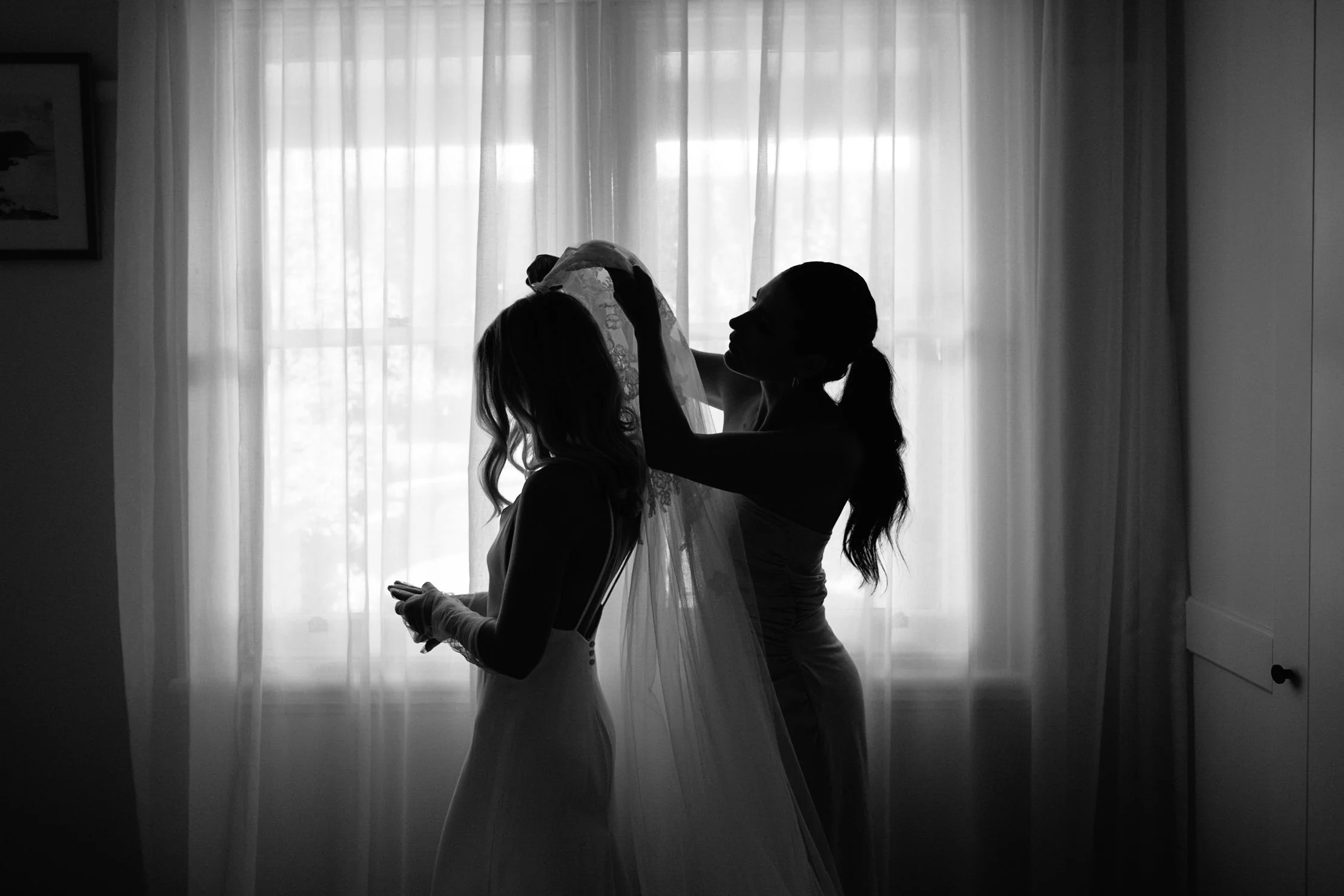 A silhouette of a bride and another woman, possibly her mother, with the bride holding a veil, standing in front of a window with sheer curtains.