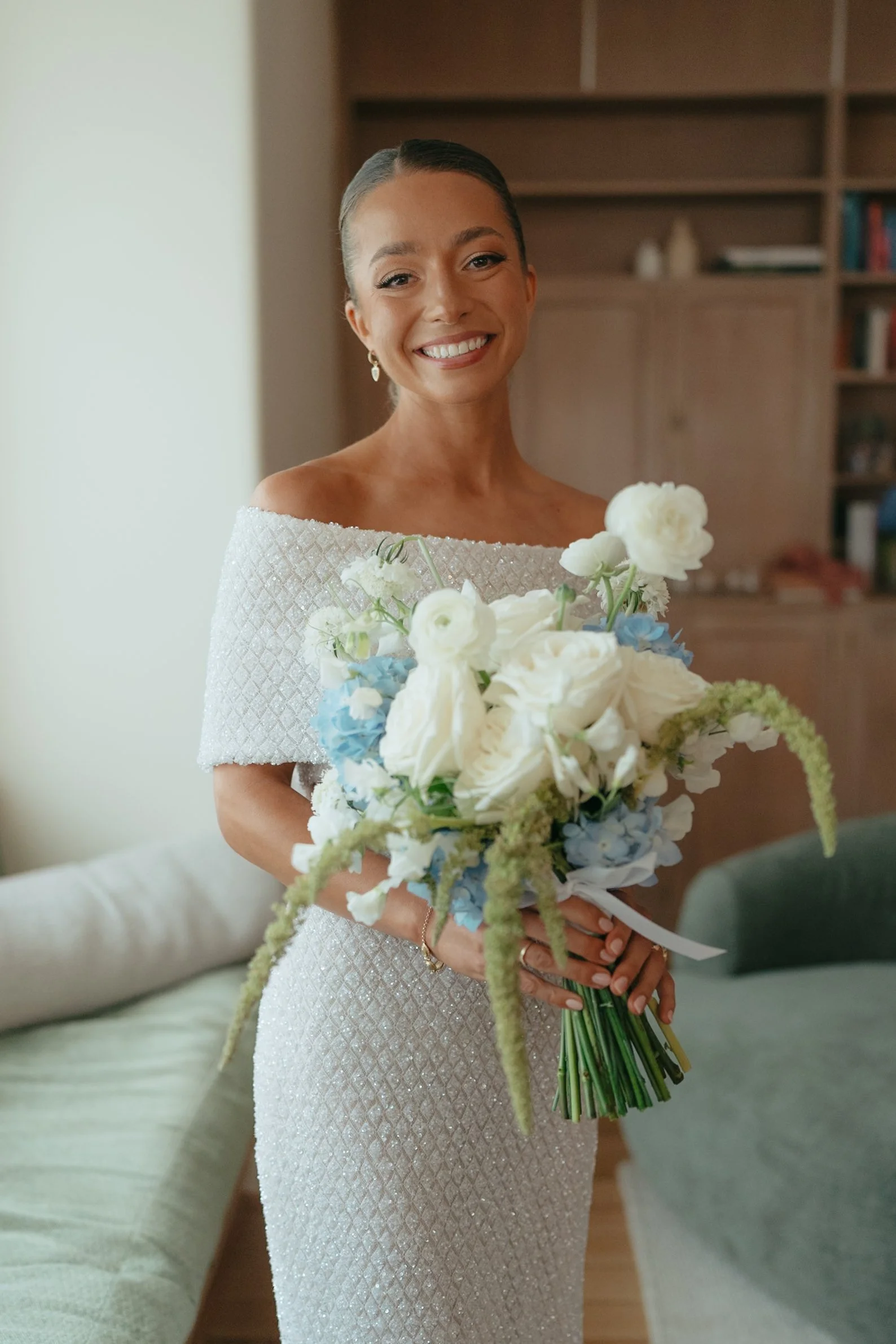 A smiling woman in a white lace wedding dress is holding a bouquet of white and blue flowers. She has her hair slicked back and is wearing pearl earrings.