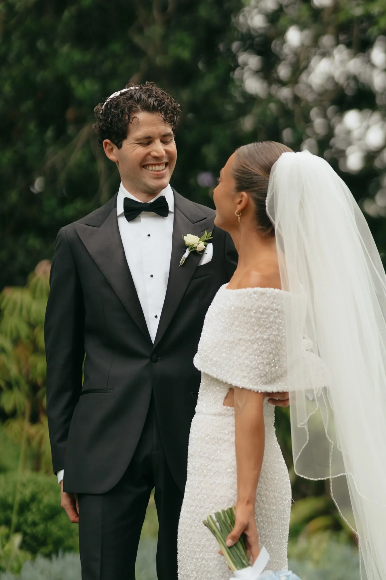 A groom in a tuxedo smiling at a bride in a white wedding dress holding a bouquet, standing outdoors in a green garden.