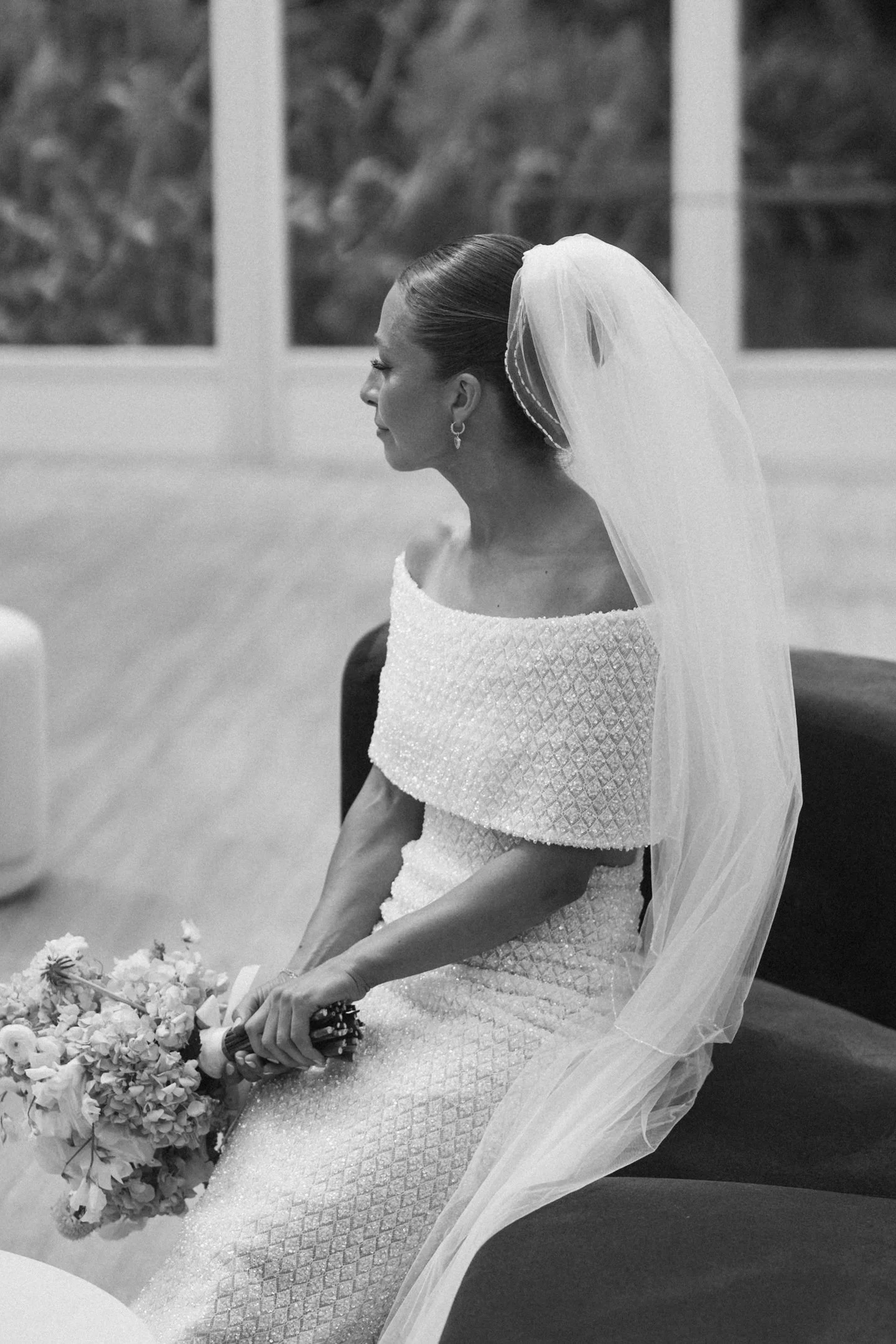 A bride with dark hair in an updo, wearing a white off-the-shoulder wedding dress with textured fabric and a veil, sits holding a bouquet of flowers in her lap at a wedding ceremony. She is wearing earrings and is in profile, with a serene expression
