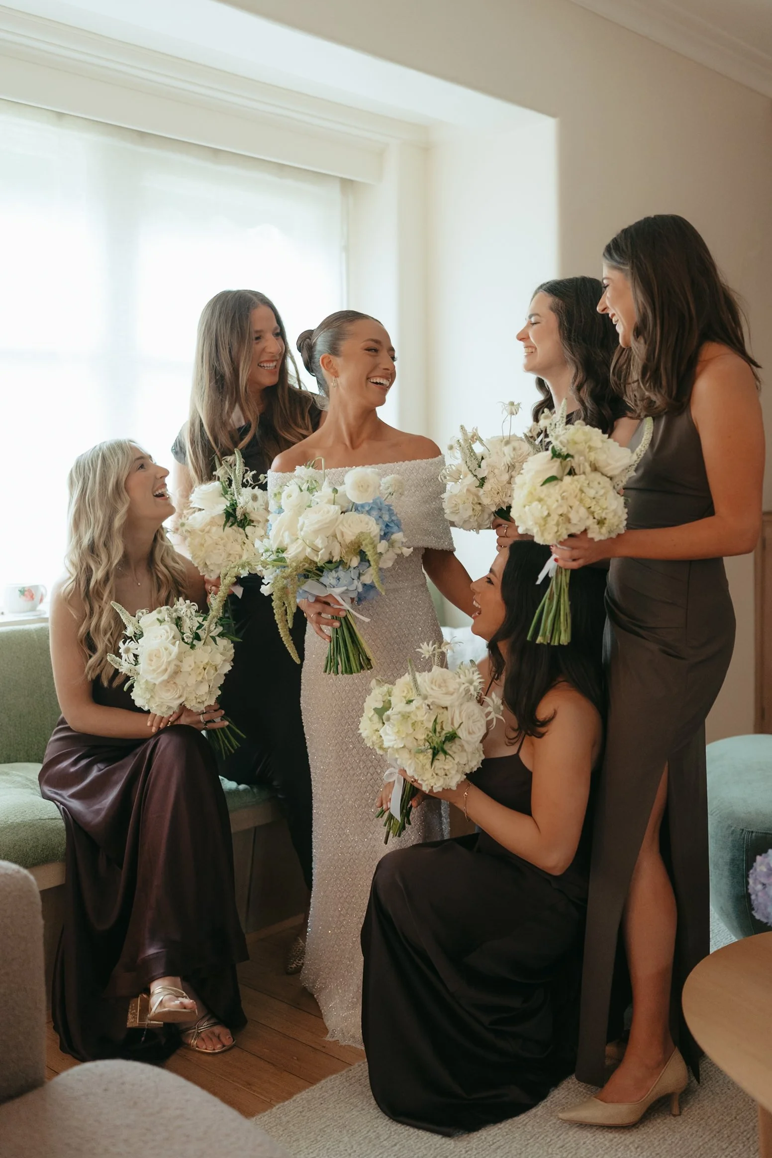 A group of women dressed in formal attire, holding bouquets, happily gathering indoors with natural light from a large window.