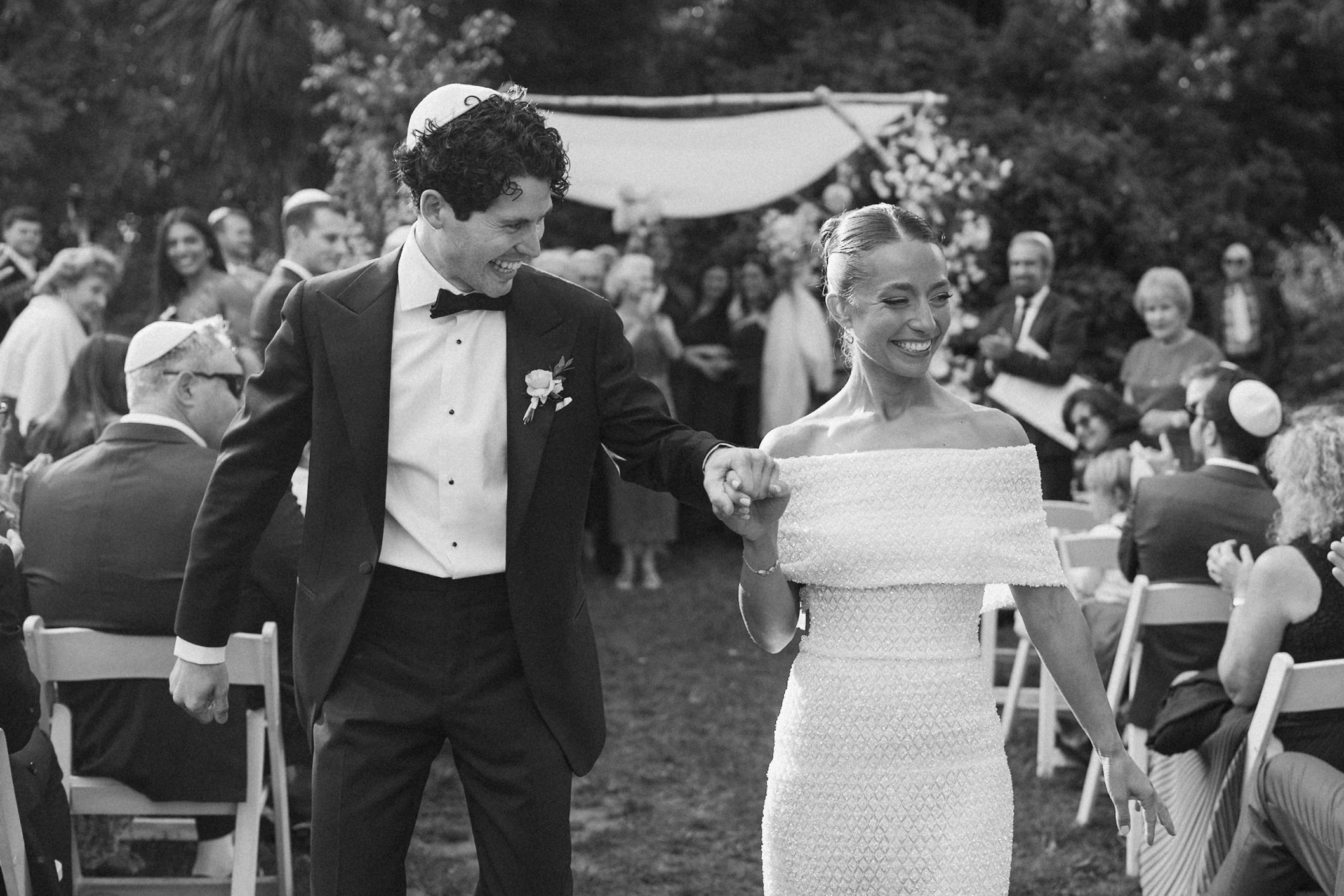 A black and white photo of a wedding ceremony outdoors. A smiling man wearing a tuxedo and bow tie holds the hand of a smiling woman in an elegant off-the-shoulder wedding dress. Guests are seated and standing in the background.