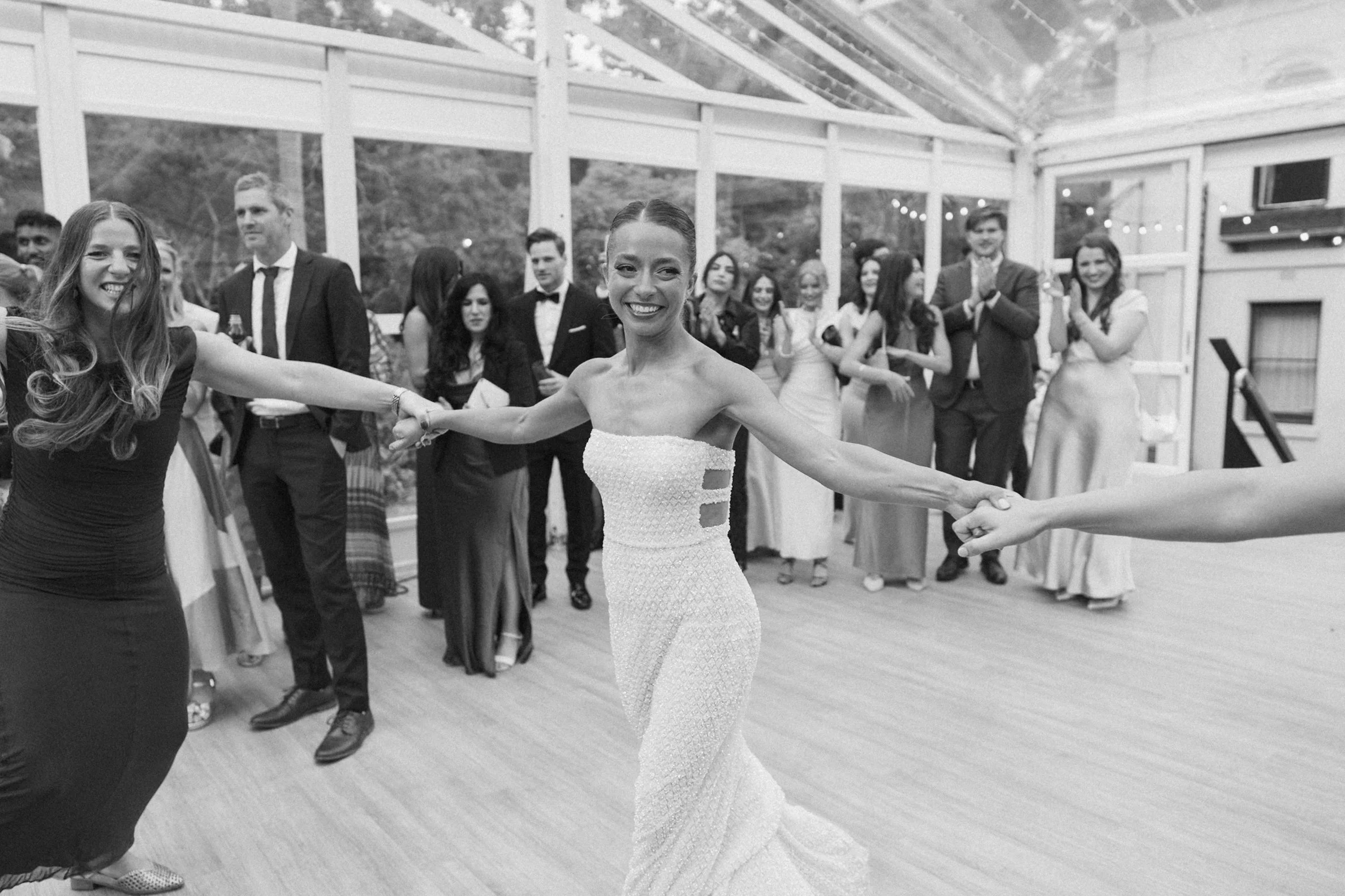 A group of people at a wedding dance, with a bride in a strapless wedding dress holding hands with a woman in a black dress, smiling in a bright, decorated venue.