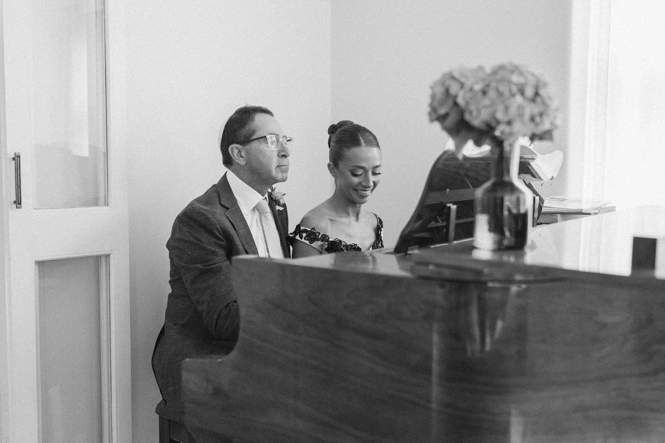 A black-and-white photo of a man and a woman playing piano together in a well-lit room, with the woman smiling and the man concentrating.