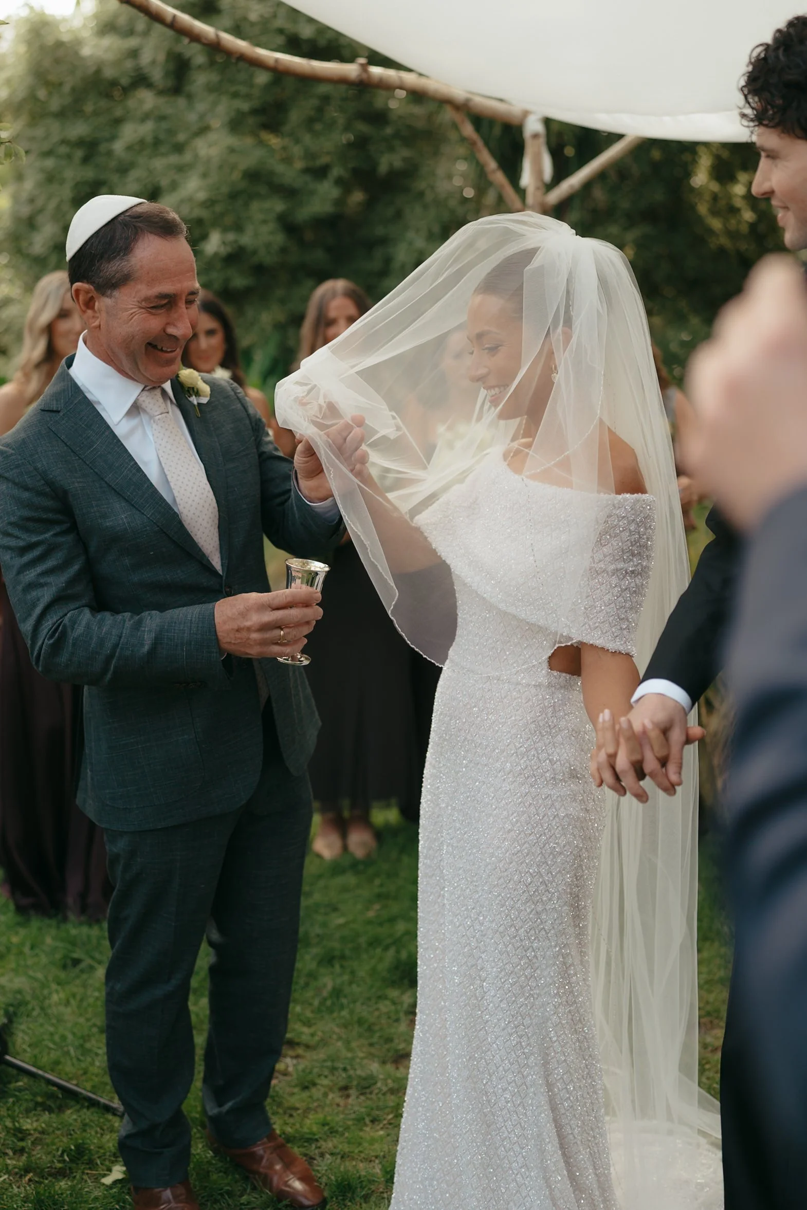 A wedding ceremony outdoors where a bride in a sparkling white dress with a veil is smiling as she holds hands with her groom. An older man, possibly the officiant, wearing a kippah and holding a small cup, is smiling and appears to be reading or spe