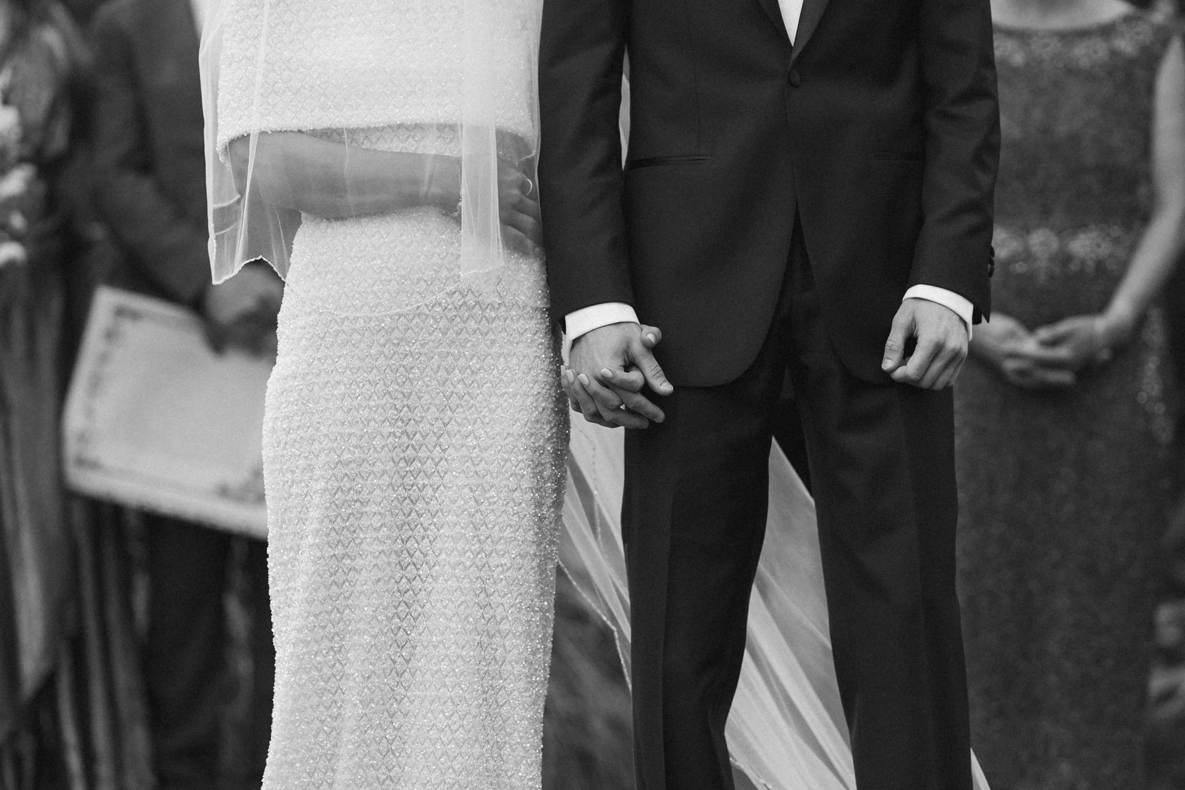 A couple holding hands during a wedding ceremony, with the bride in a white dress and the groom in a dark suit, standing outdoors.