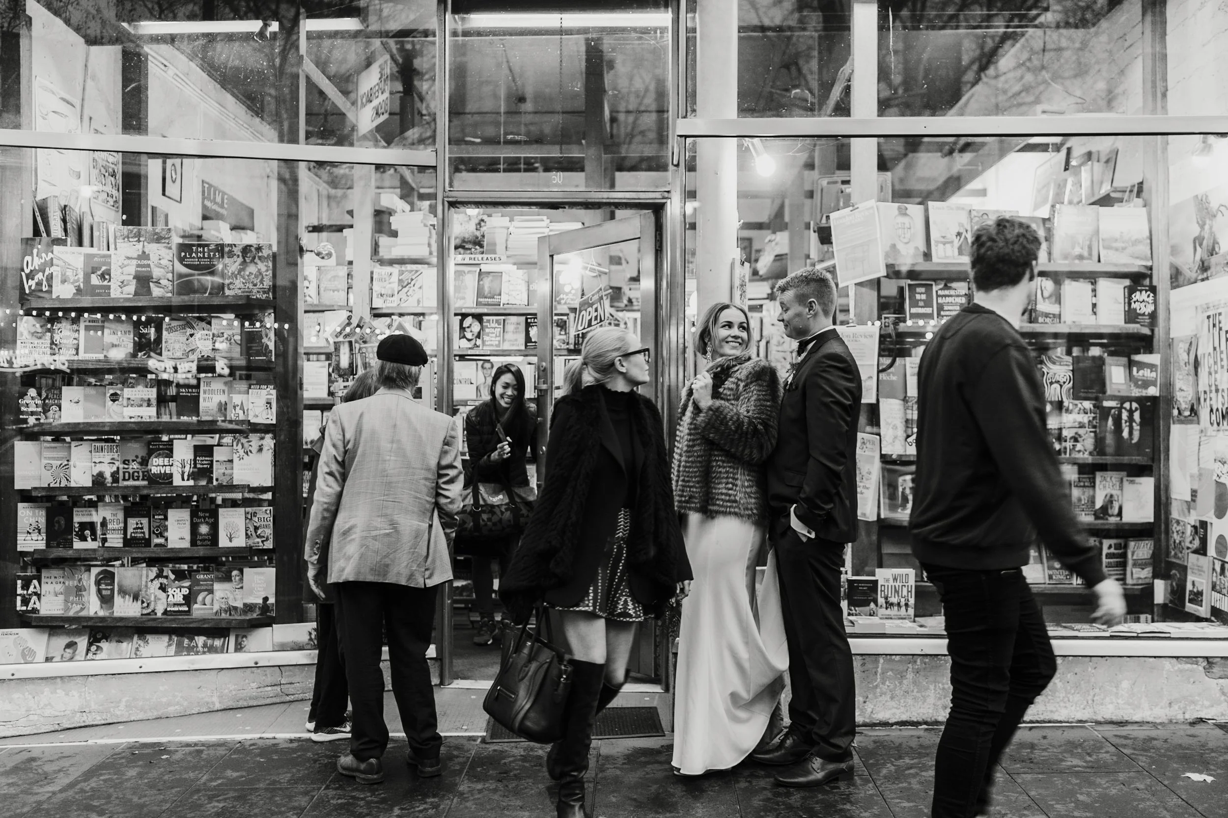 Black and white photo of a bookstore with several people walking and talking outside, the storefront has glass windows filled with books and signs, including an 