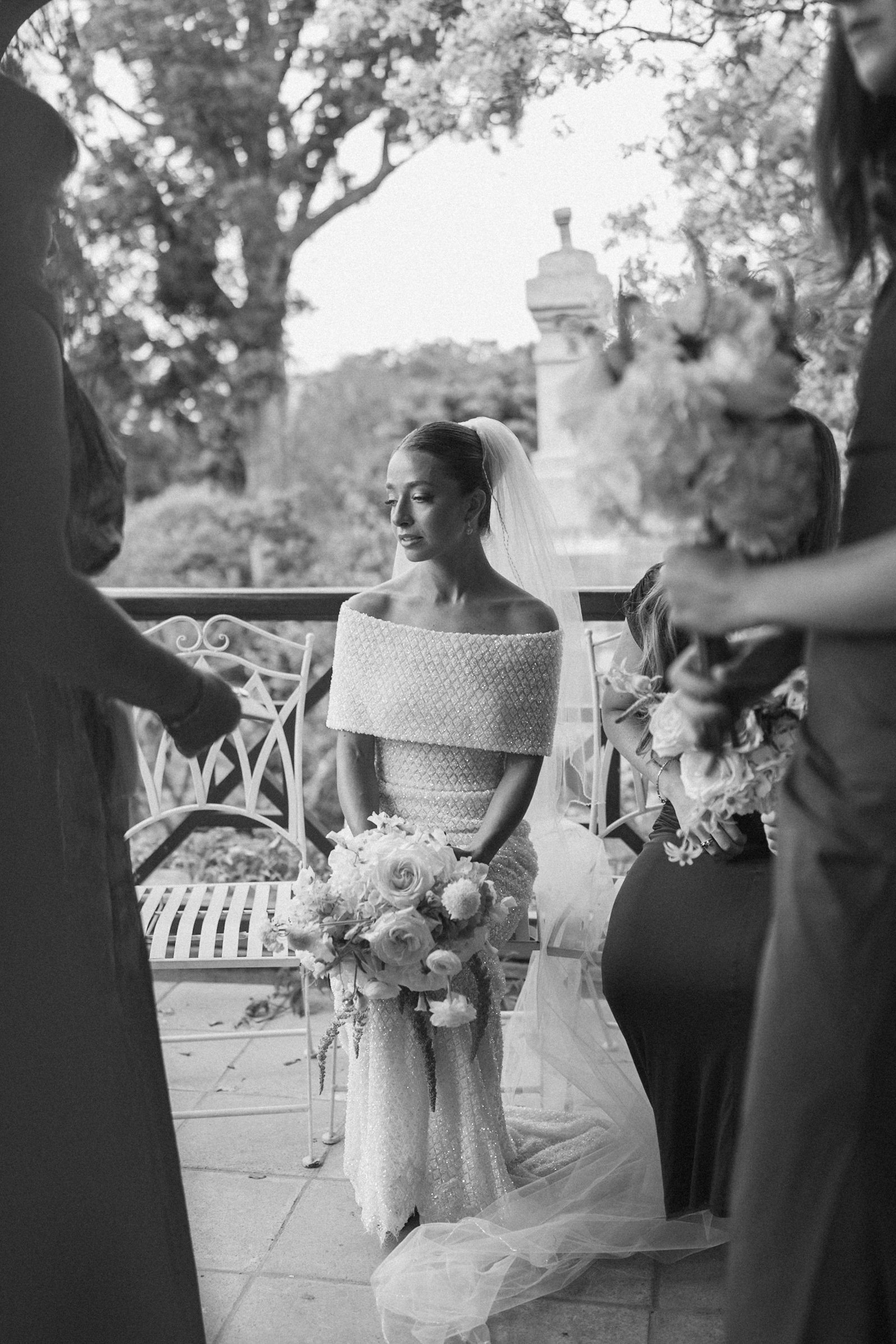 A bride in a wedding dress with a veil holding a bouquet of flowers, during a wedding ceremony outdoors with trees and a stone structure in the background.