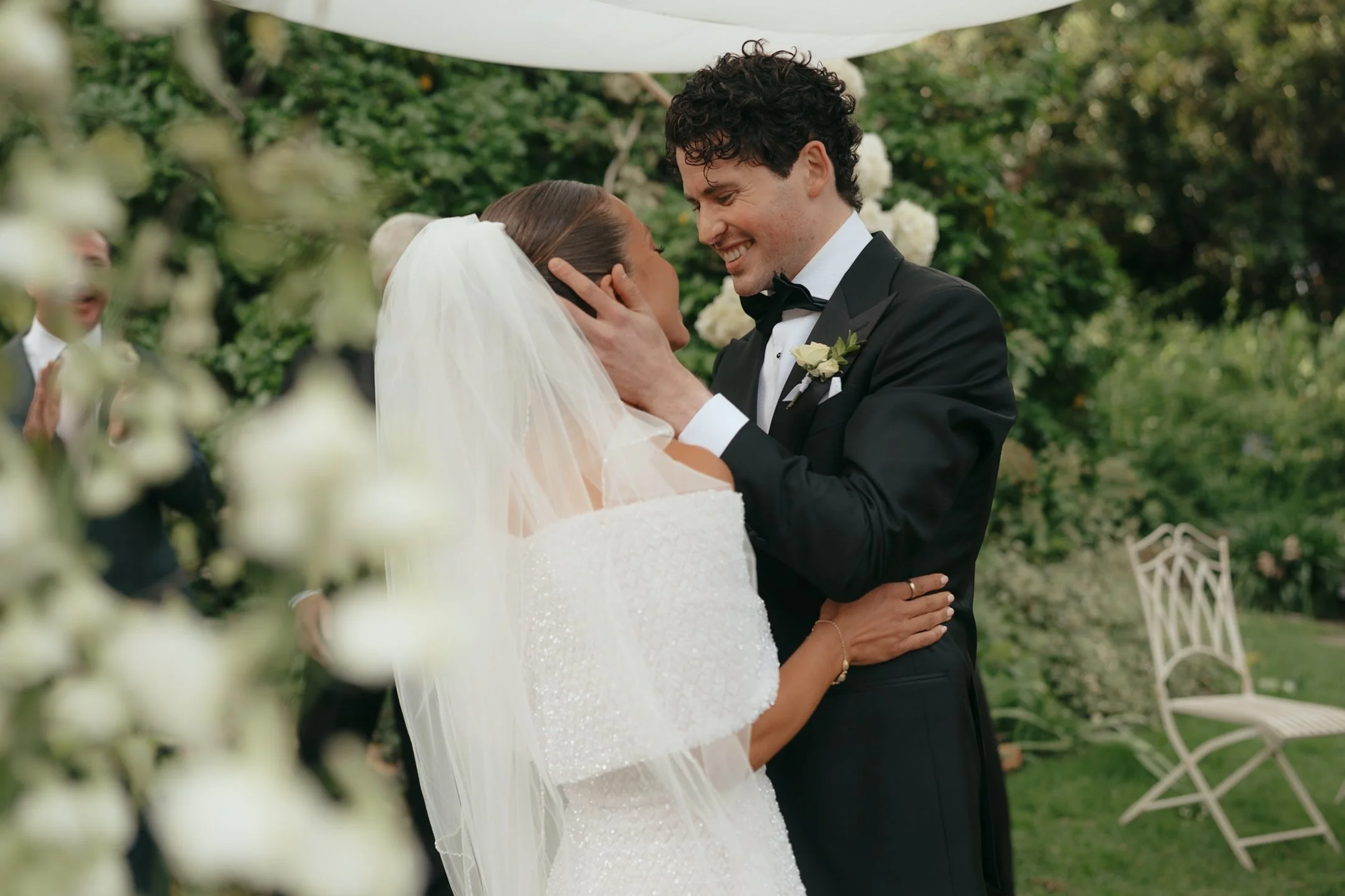 A bride and groom share a joyful moment during their outdoor wedding ceremony, with the bride in a white dress and veil, and the groom in a black tuxedo, surrounded by greenery and floral decorations.