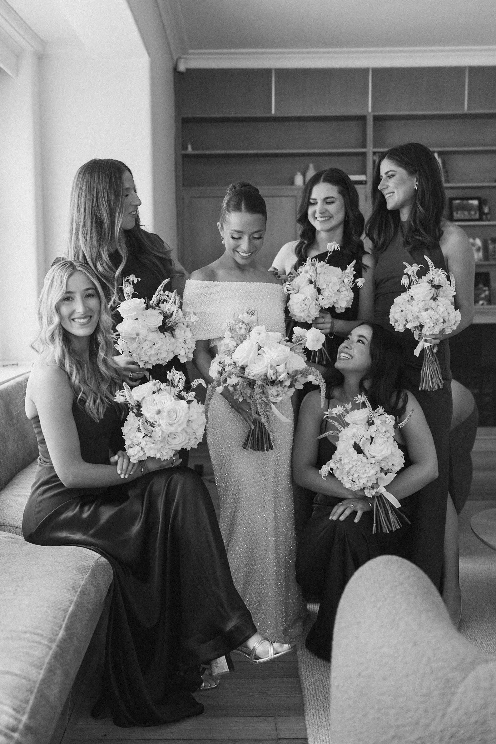 Black and white photo of five women dressed in formal attire holding bouquets, gathered in a room with shelves in the background. The women are smiling, some looking at each other or down at their bouquets.