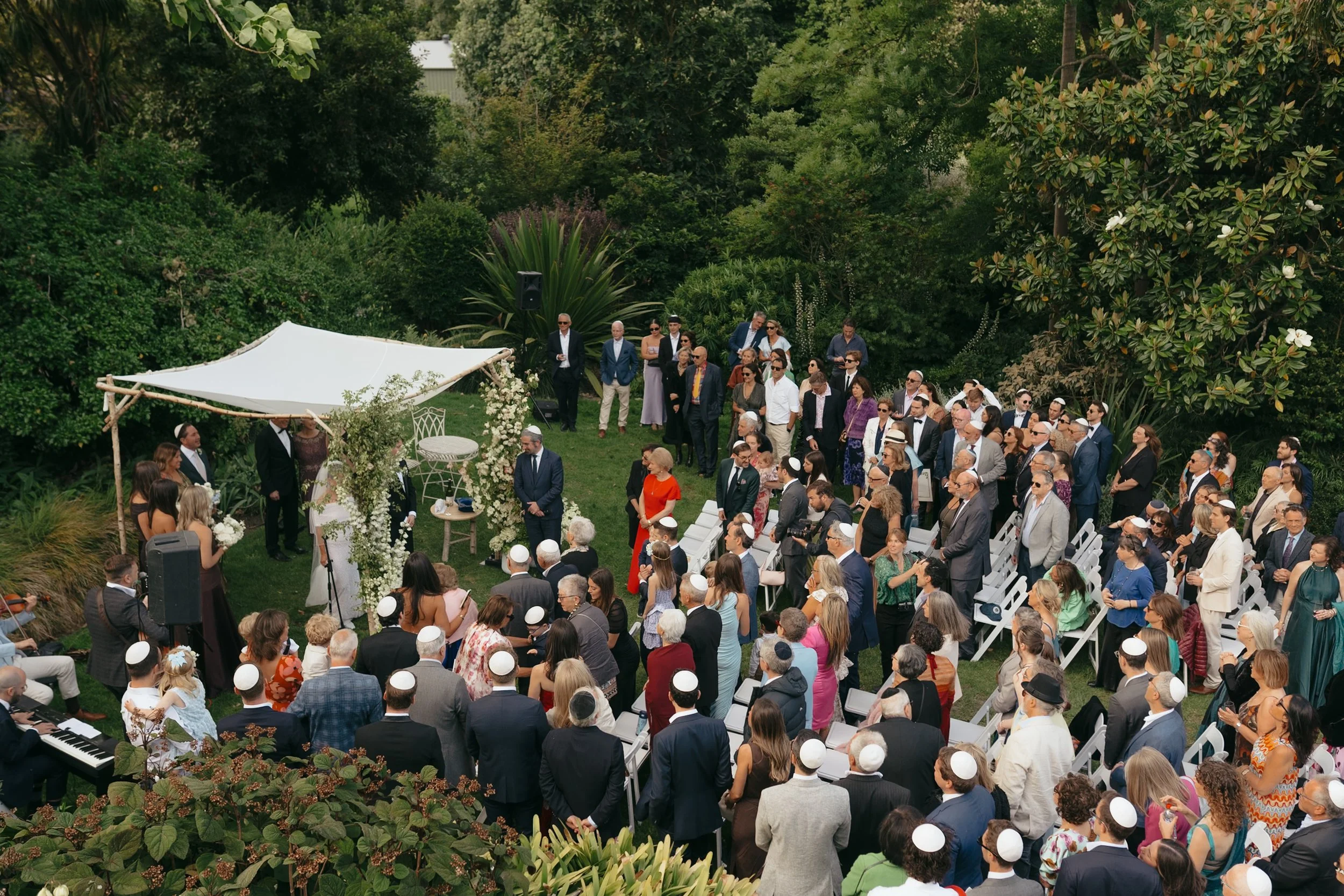 Outdoor wedding ceremony with a large gathering of guests, some wearing yarmulkes, in a lush garden setting with greenery and trees.