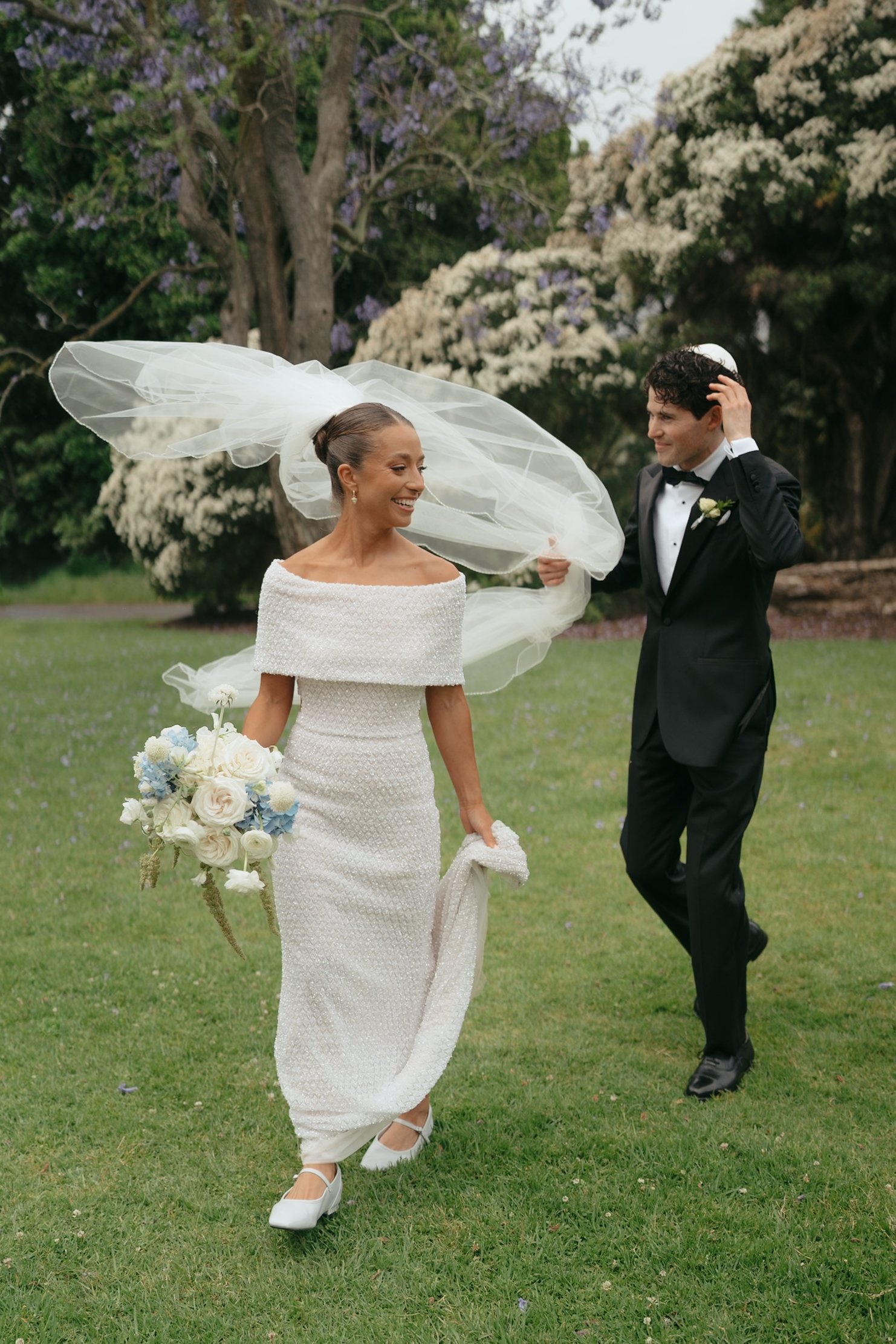 A bride in a white wedding dress holding a bouquet of white and blue flowers, standing outdoors with a man in a black tuxedo who is smiling and adjusting a white kippah on his head, in a park with green grass and trees with white and purple blossoms.