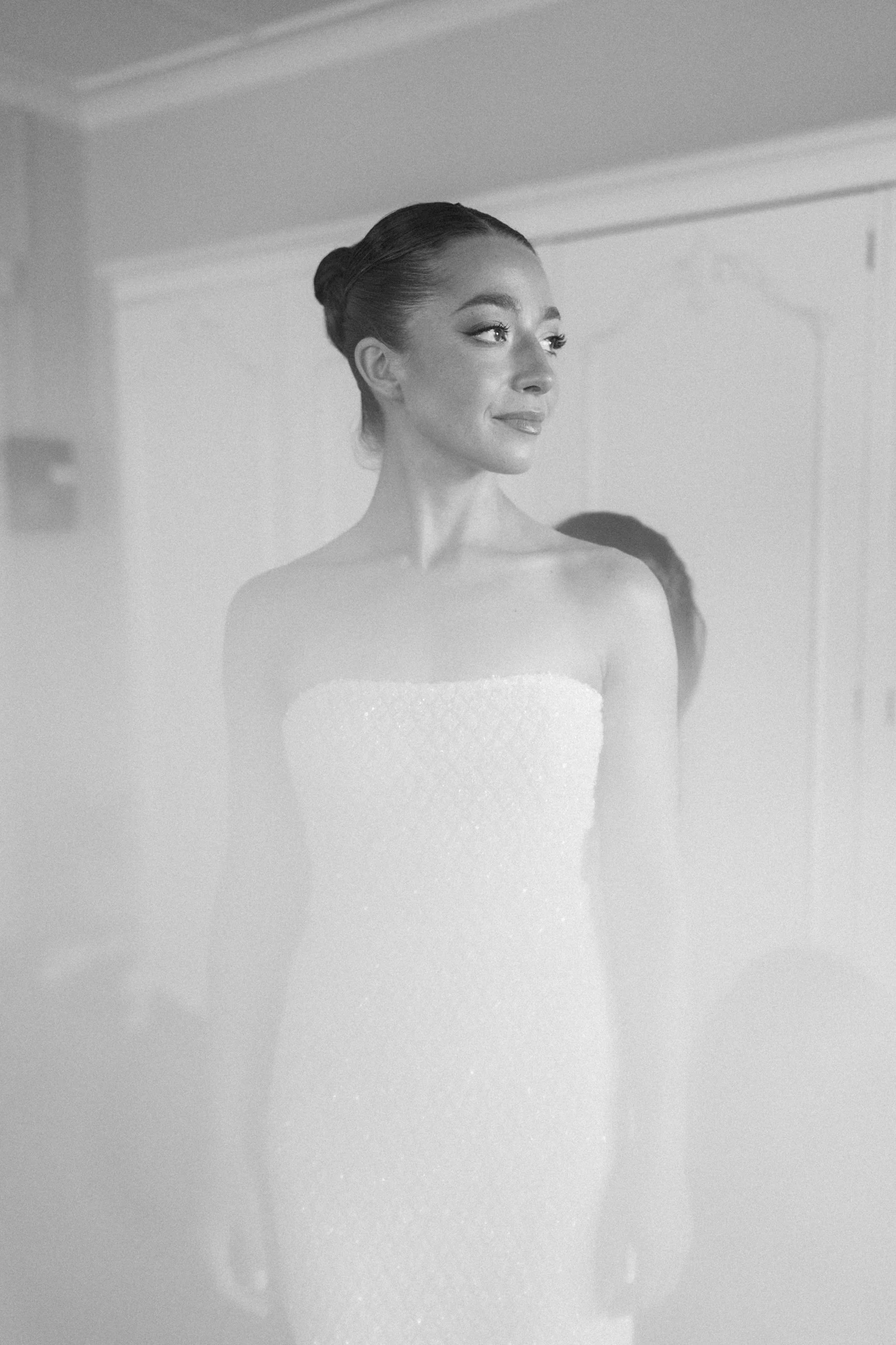 A woman with makeup and styled hair, smiling slightly, wearing a strapless dress or top, standing in a room with a blurred background.