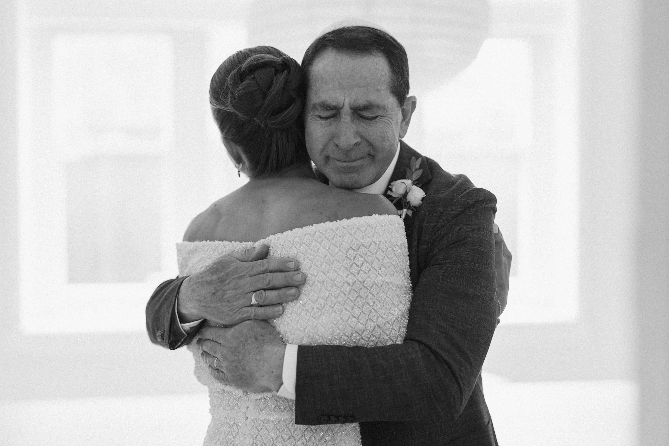 A black and white photo of a man in a suit and a woman in a wedding dress hugging tightly, with their eyes closed, expressing emotion.