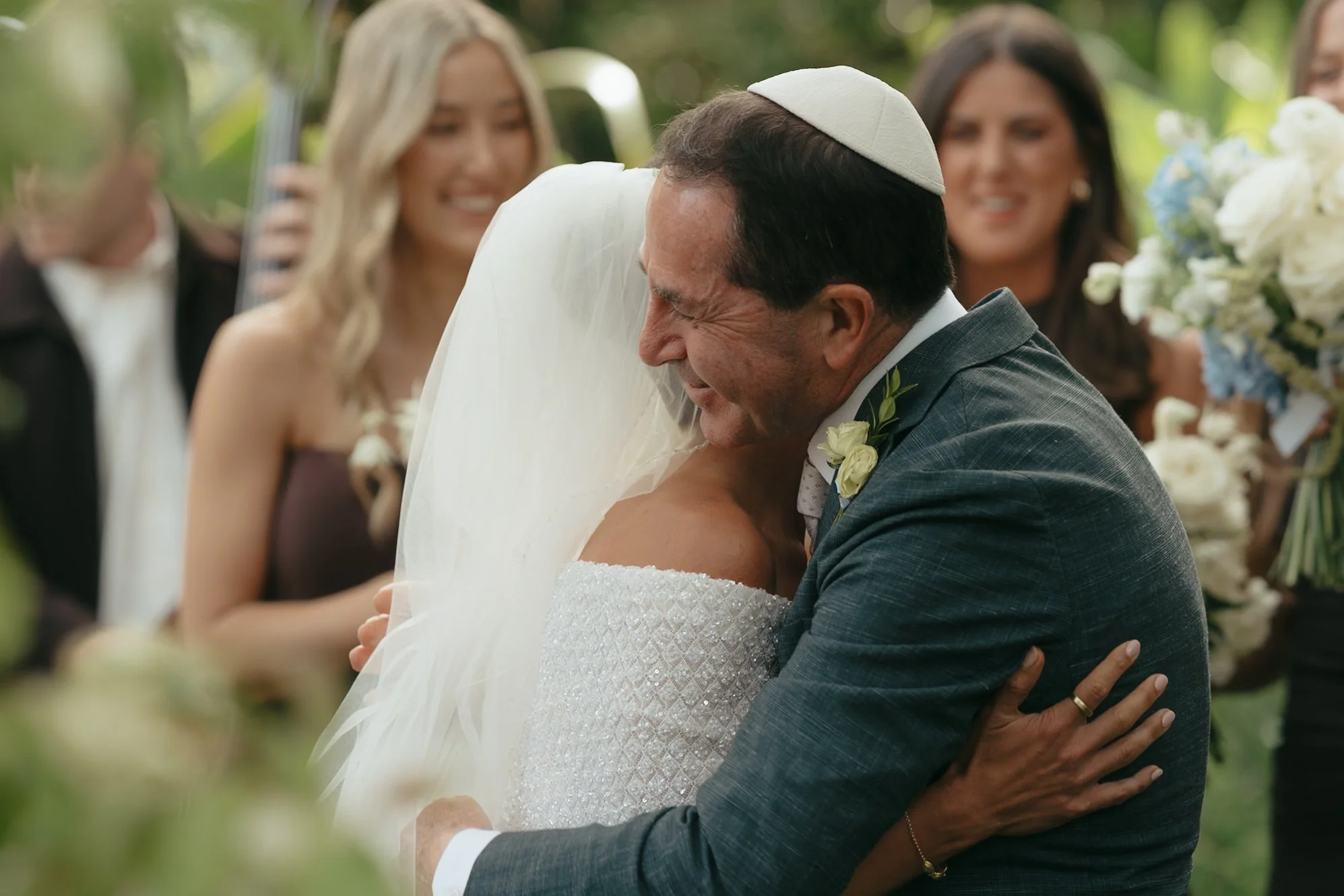 A bride in a white wedding dress and veil hugging an older man wearing a gray suit and a white kippah, smiling amid a crowd of wedding guests in an outdoor setting.