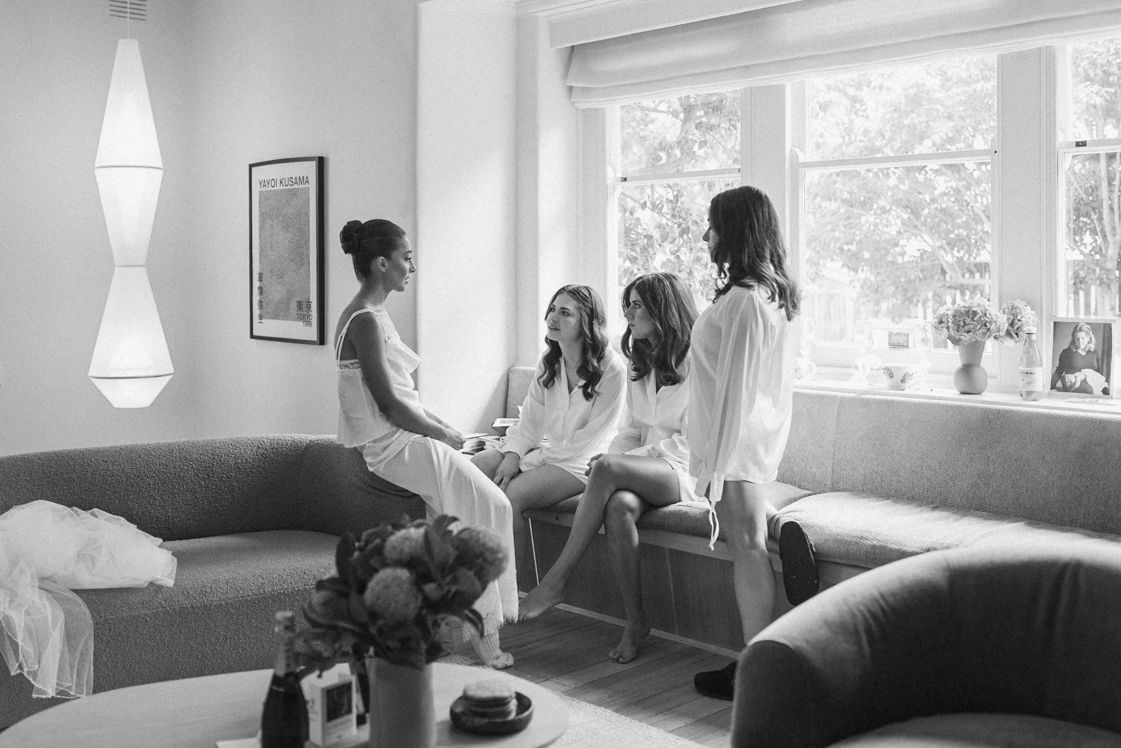 Four women in white robes in a bright living room, two seated on a couch and two standing, engaged in conversation.