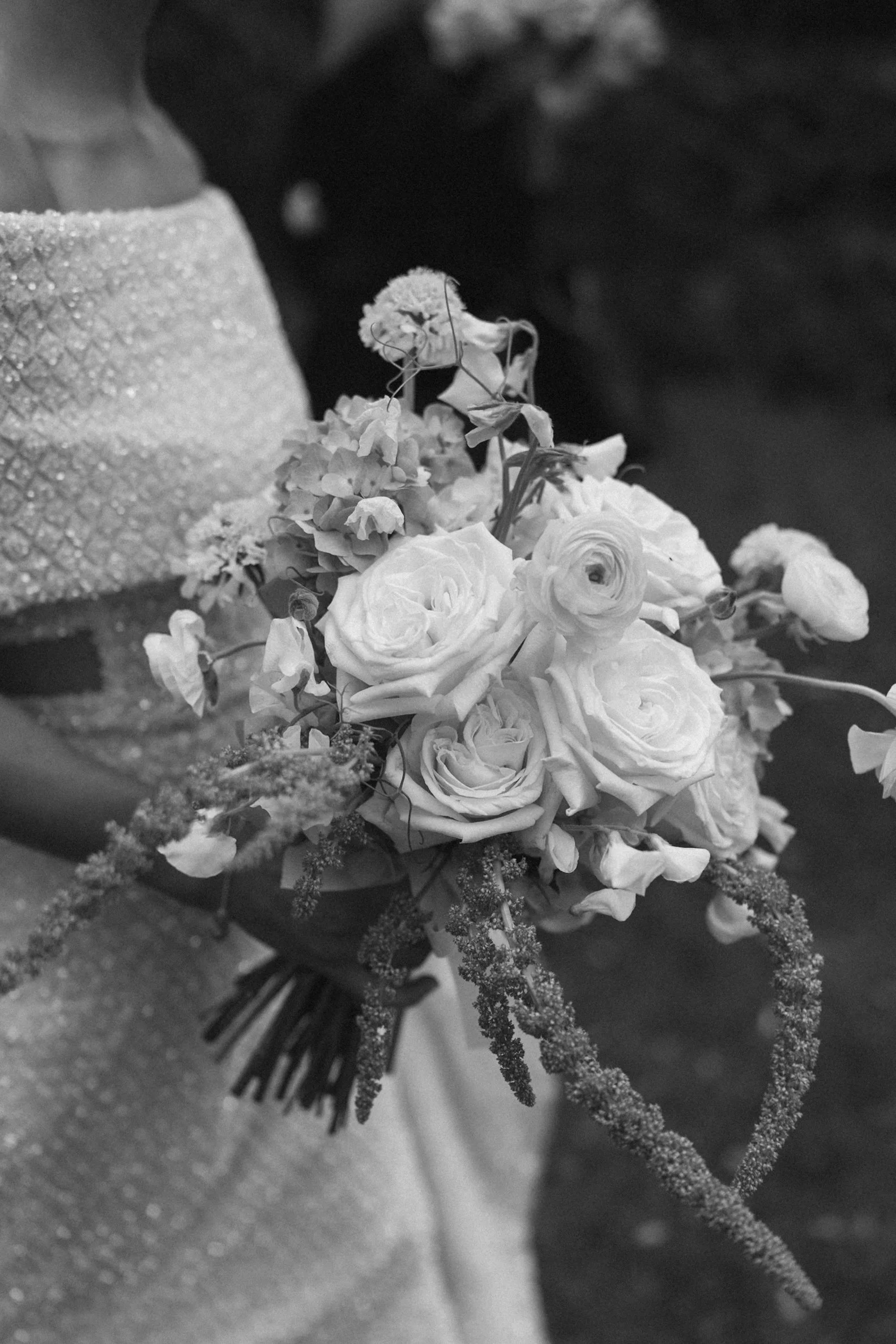 Black and white photo of a bouquet of flowers, including roses, with a sparkly hat or headpiece in the background.
