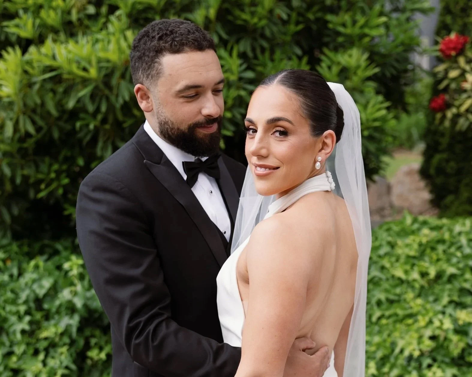 A bride and groom in wedding attire embracing outdoors, with green foliage in the background. The groom is in a black tuxedo with a bow tie, and the bride is in a white dress with a veil and pearl earrings.
