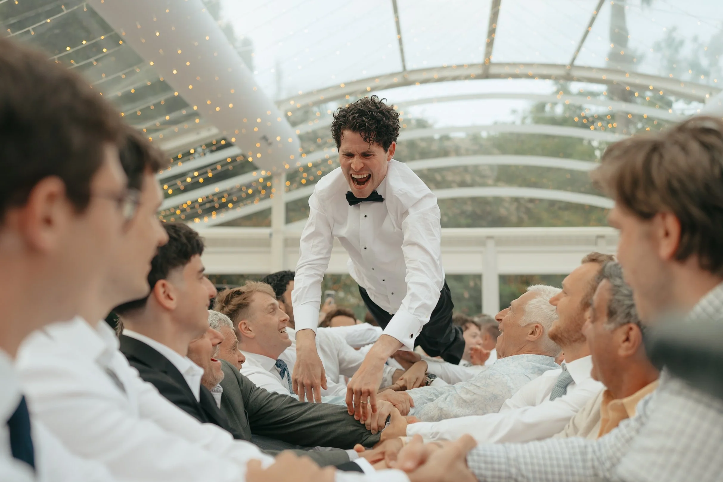 A man in a white shirt and black bow tie is standing and laughing on a decorated rooftop with string lights, surrounded by seated people in formal attire holding hands or forming a human chain.