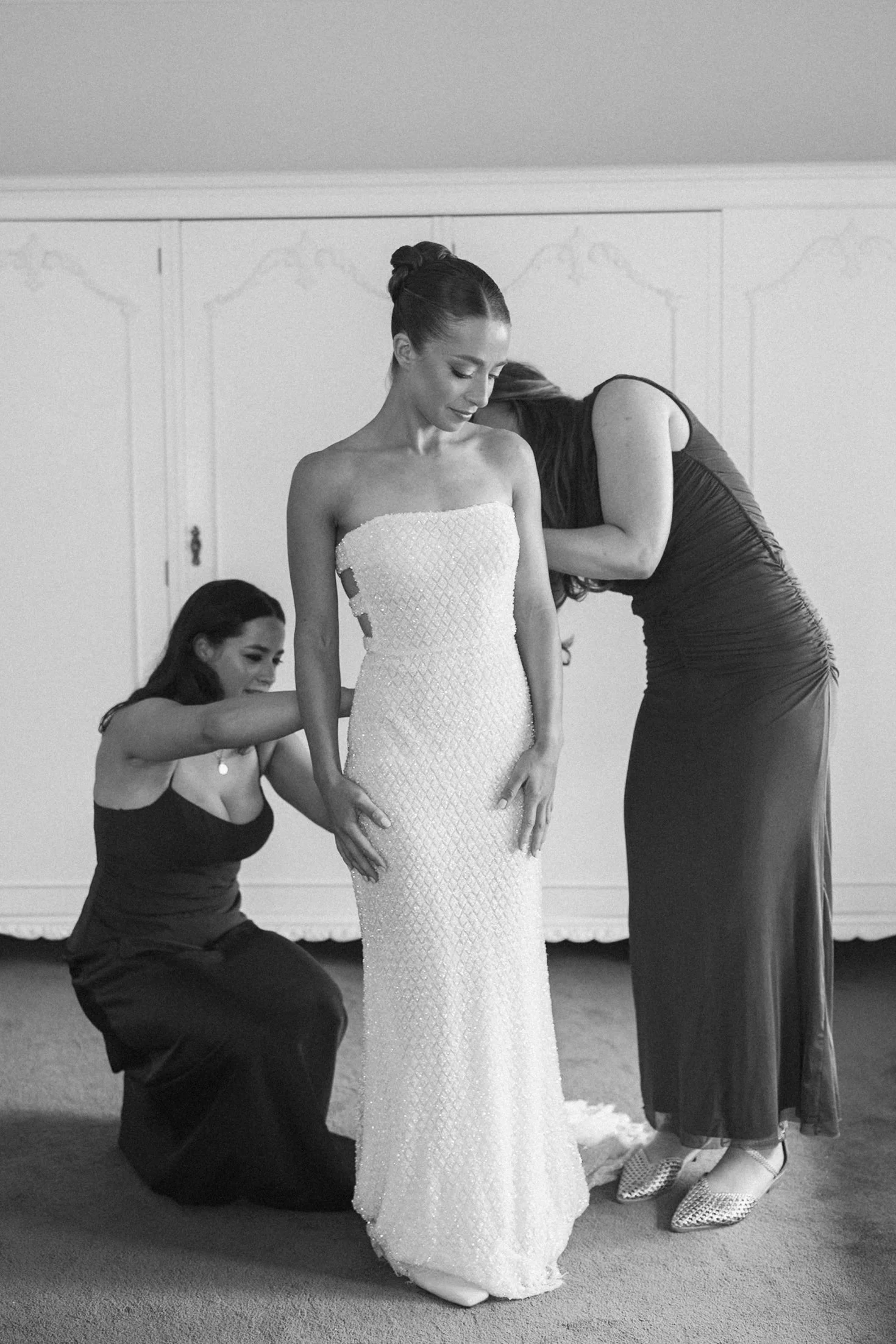 A bride in a strapless wedding gown being assisted by three women, one kneeling and two standing, in a room with plain walls.