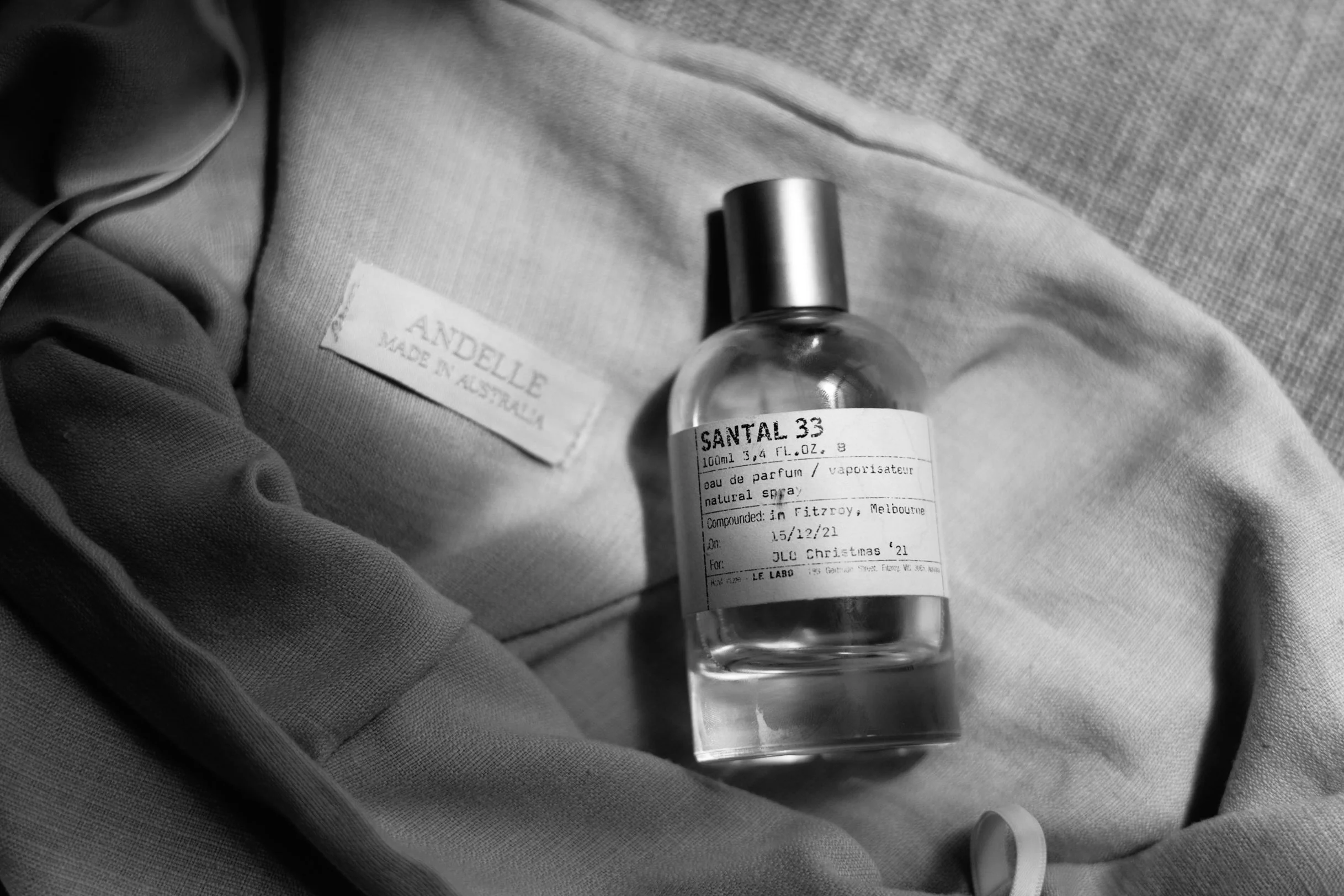 A small transparent perfume bottle labeled 'Santal 33' resting on a fabric surface, with a clothing tag that reads 'Andelle Made in Australia' nearby.