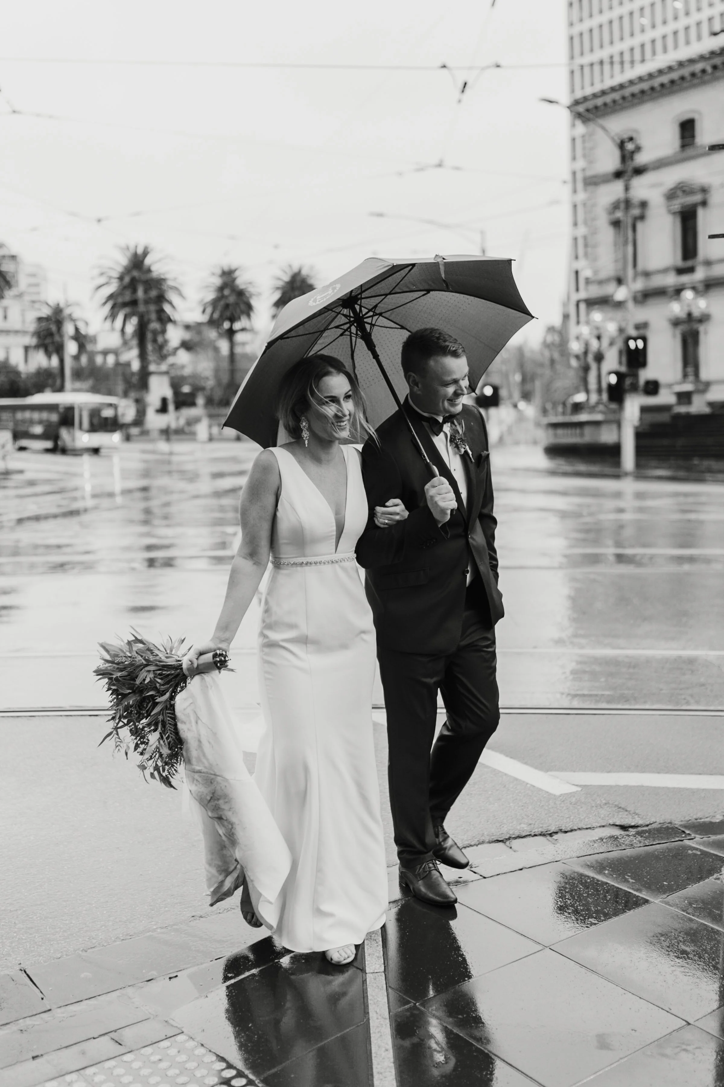 A bride and groom walking arm in arm in the rain, sharing an umbrella. The bride is wearing a white wedding dress and holding a bouquet, while the groom is in a dark tuxedo. They are smiling and walking on a city street with wet pavement, surrounded 