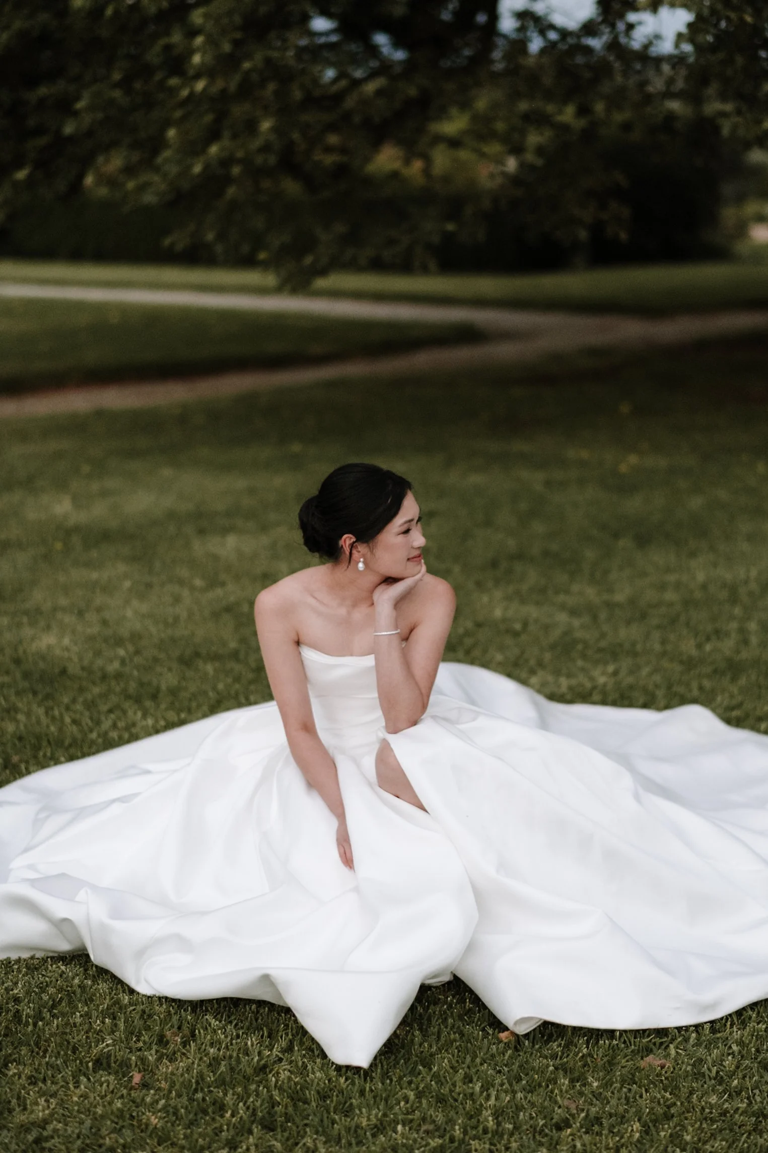 A woman in a white wedding dress sitting on the grass in a park, with trees in the background, during dusk or evening.