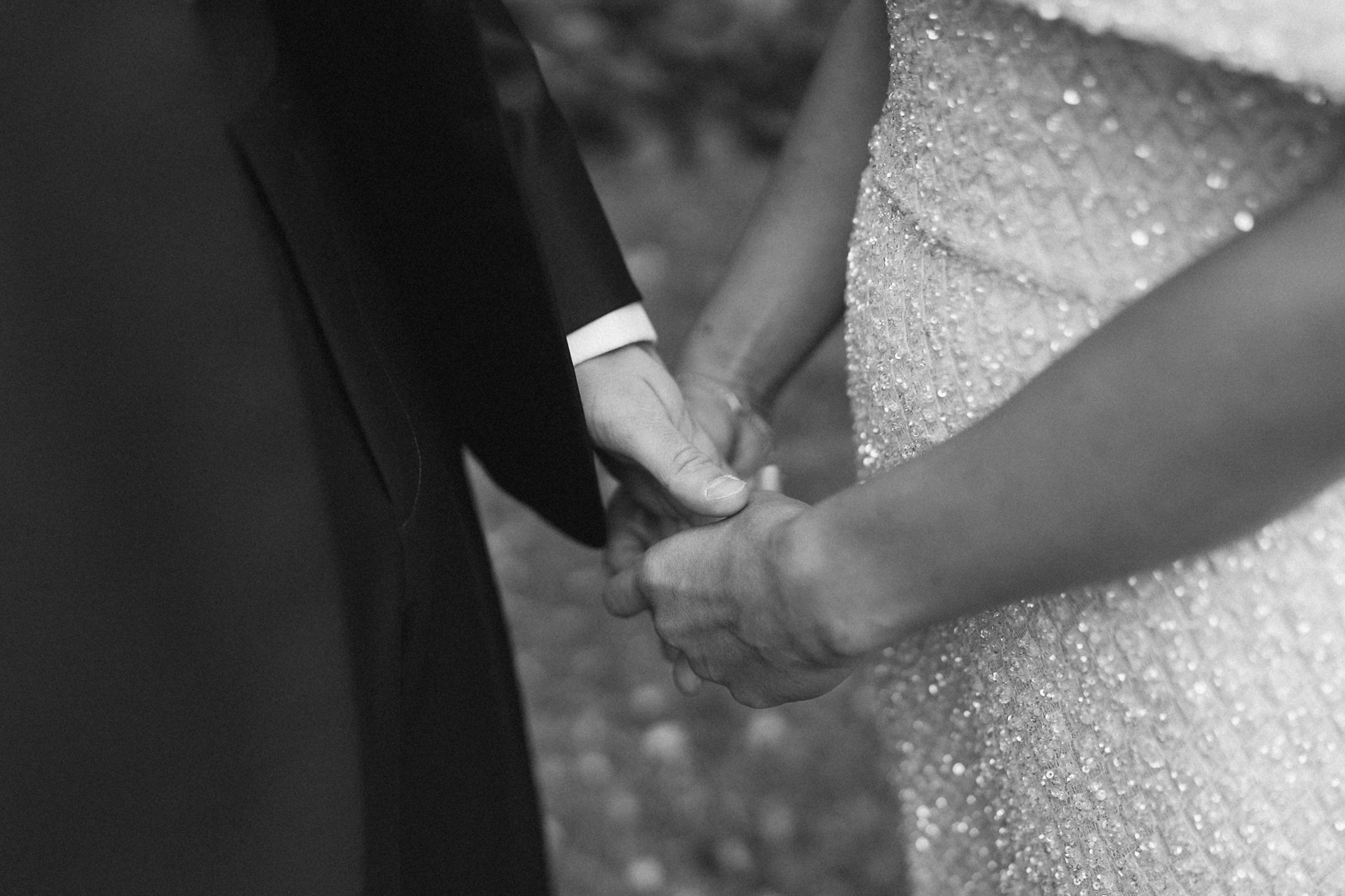 Close-up of two people holding hands, one wearing a suit, against a textured glittery surface, in black and white.