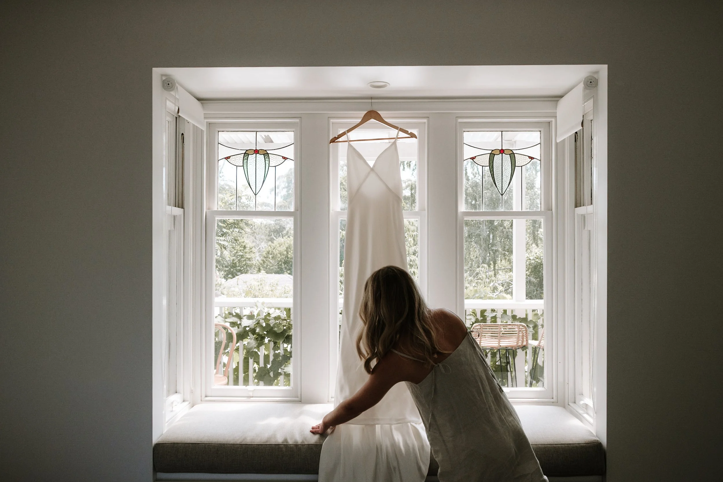 A woman kneeling on a window seat, hanging a white dress on a wooden hanger in front of a large window with stained glass accents, greenery outside.