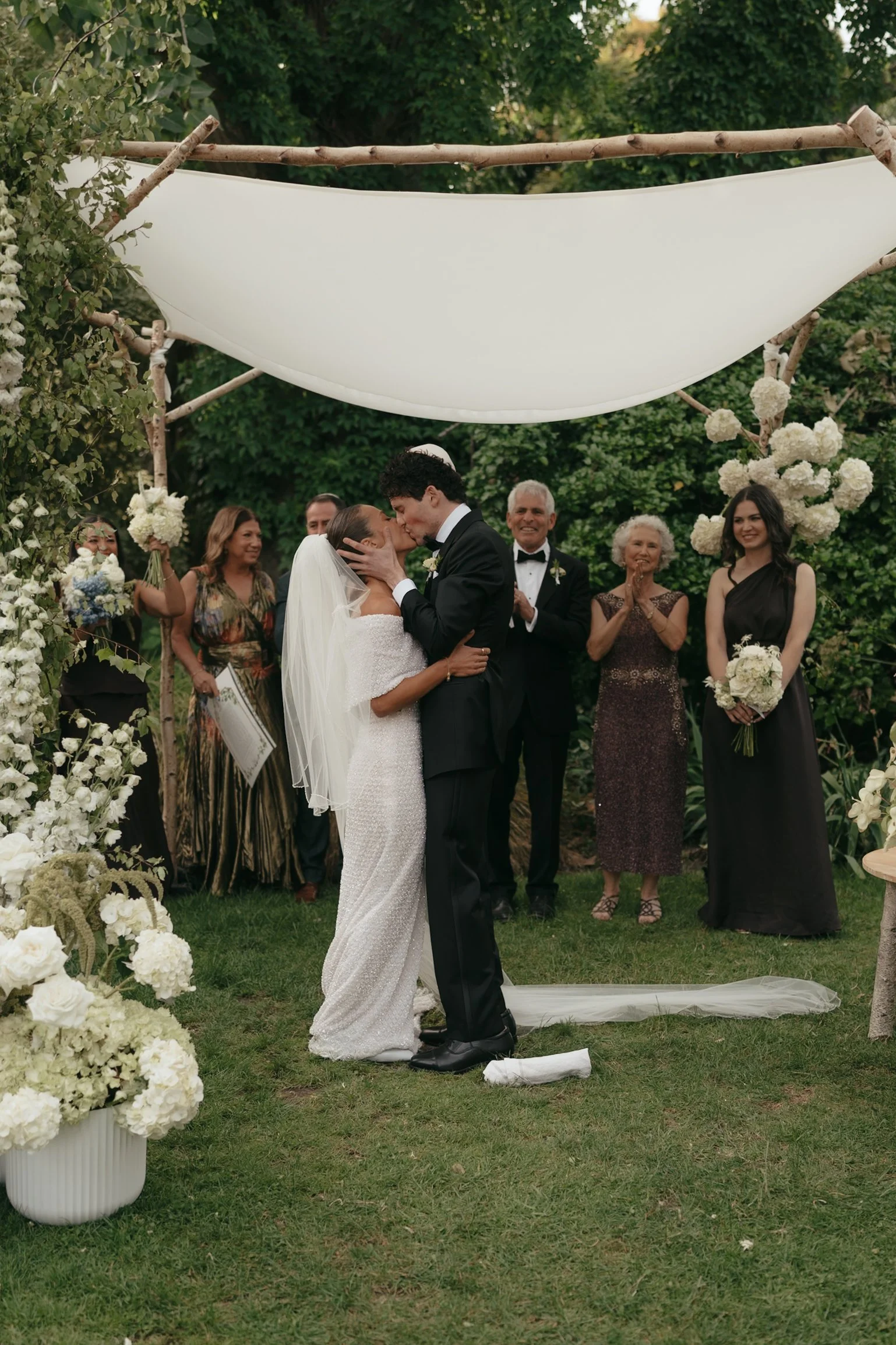 A couple getting married, kissing, during an outdoor wedding ceremony, with gathered guests standing behind them, surrounded by greenery and floral decorations.