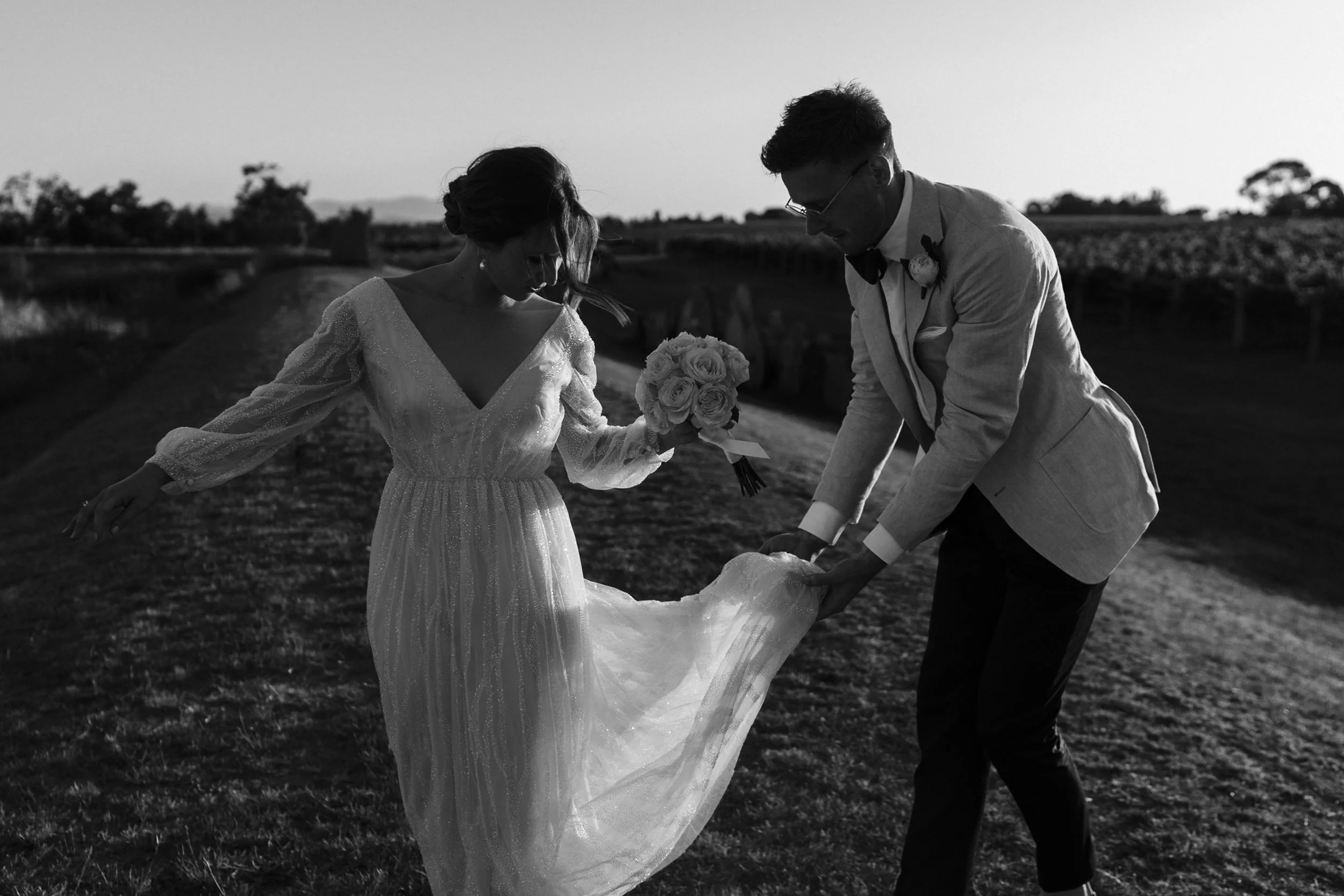 Black and white photo of a bride and groom on their wedding day outdoors. The bride is holding a bouquet of roses, and the groom is dressed in a suit with a bow tie and boutonniere. They are bending down, possibly to pick up or look at something on t