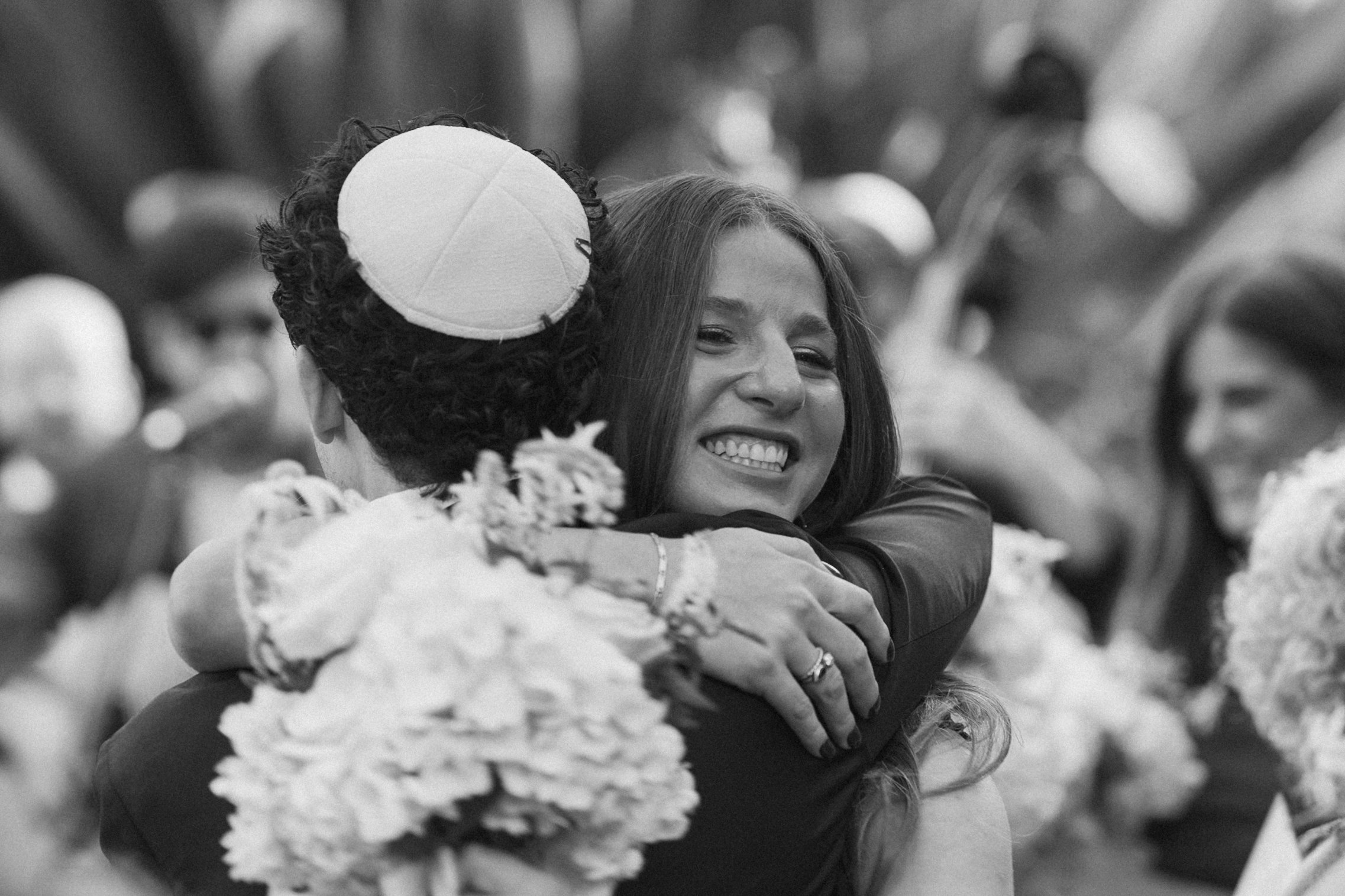 A woman with long hair happily hugging a person wearing a hat, in an outdoor setting with blurred crowd in the background.