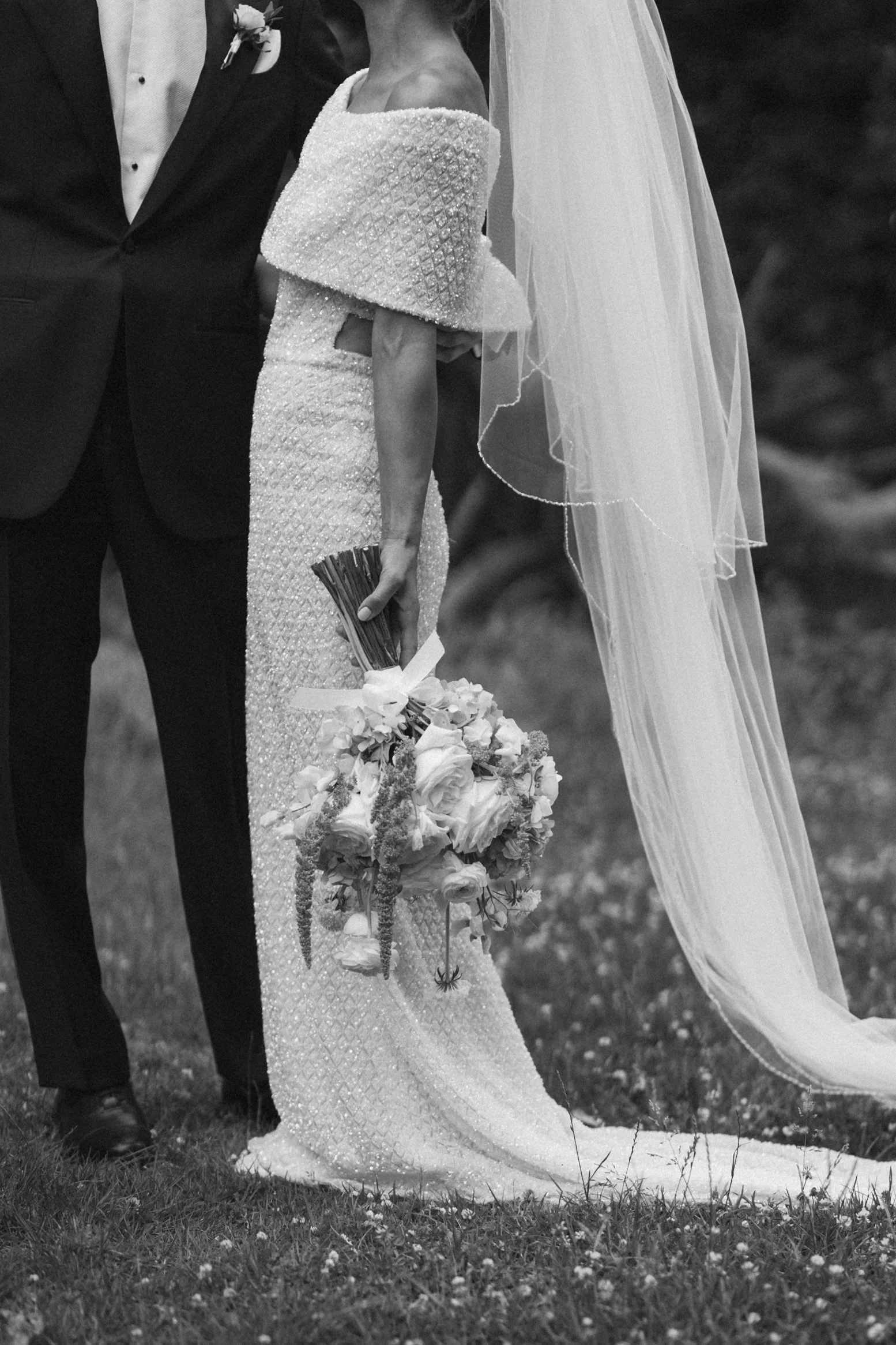 A black and white photo of a bride and groom holding hands outdoors. The bride is wearing a textured, off-the-shoulder wedding gown and veil, and holding a bouquet of flowers. The groom is dressed in a suit and tie.