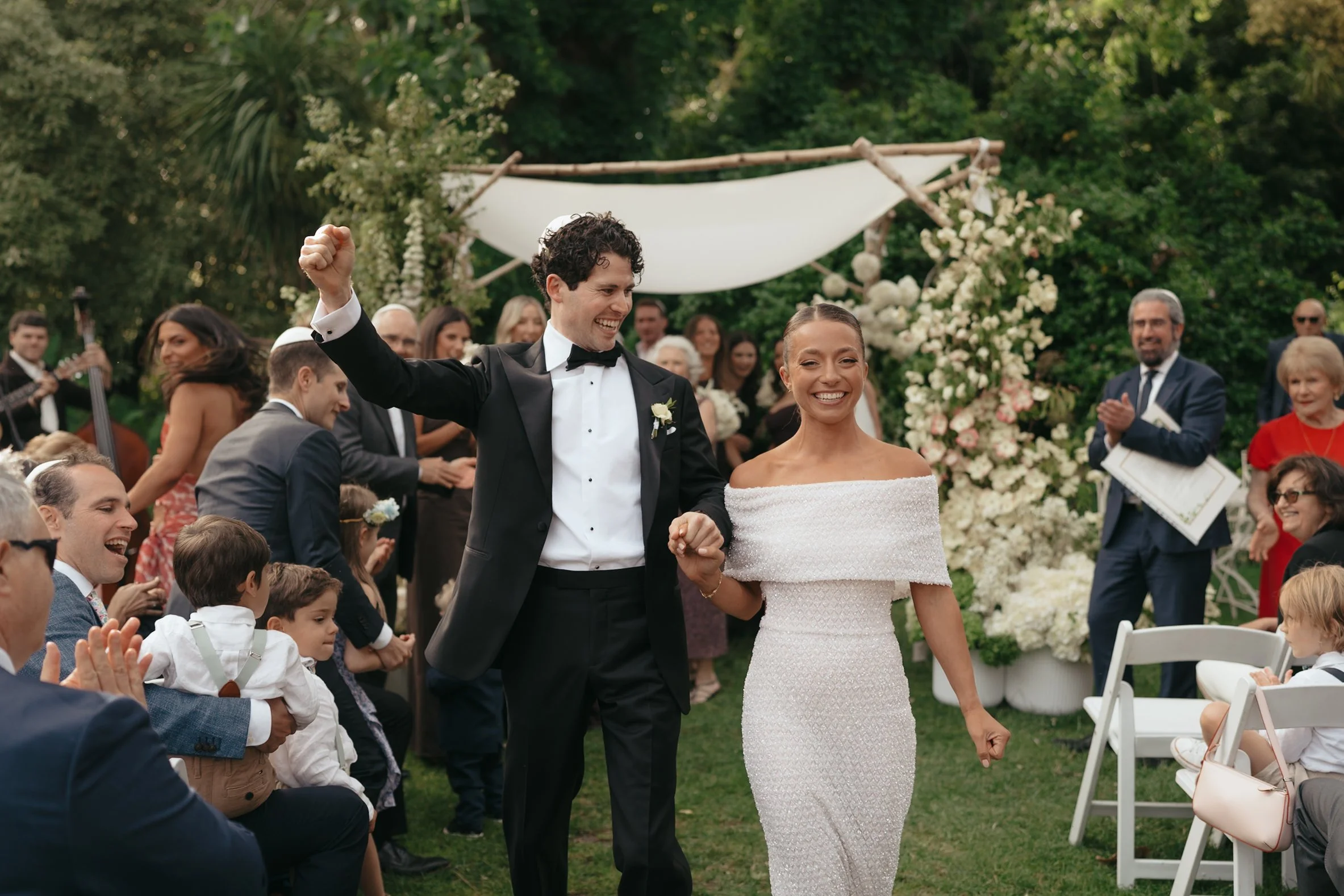 A newlywed couple walking hand in hand at their outdoor wedding ceremony, surrounded by guests and a floral backdrop.