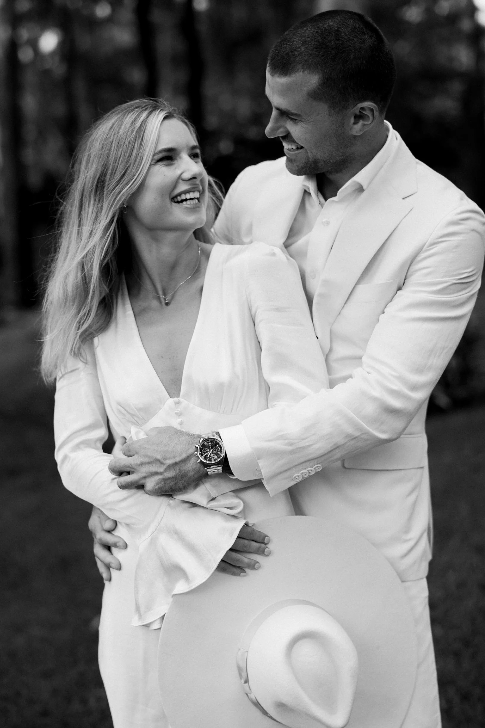 A black and white photo of a couple smiling and looking at each other. The man, wearing a white suit jacket and shirt, is holding a hat and has a watch on his left wrist. The woman is wearing a white dress and a necklace, with her hair loose. They are outdoors, with a blurred background of trees.