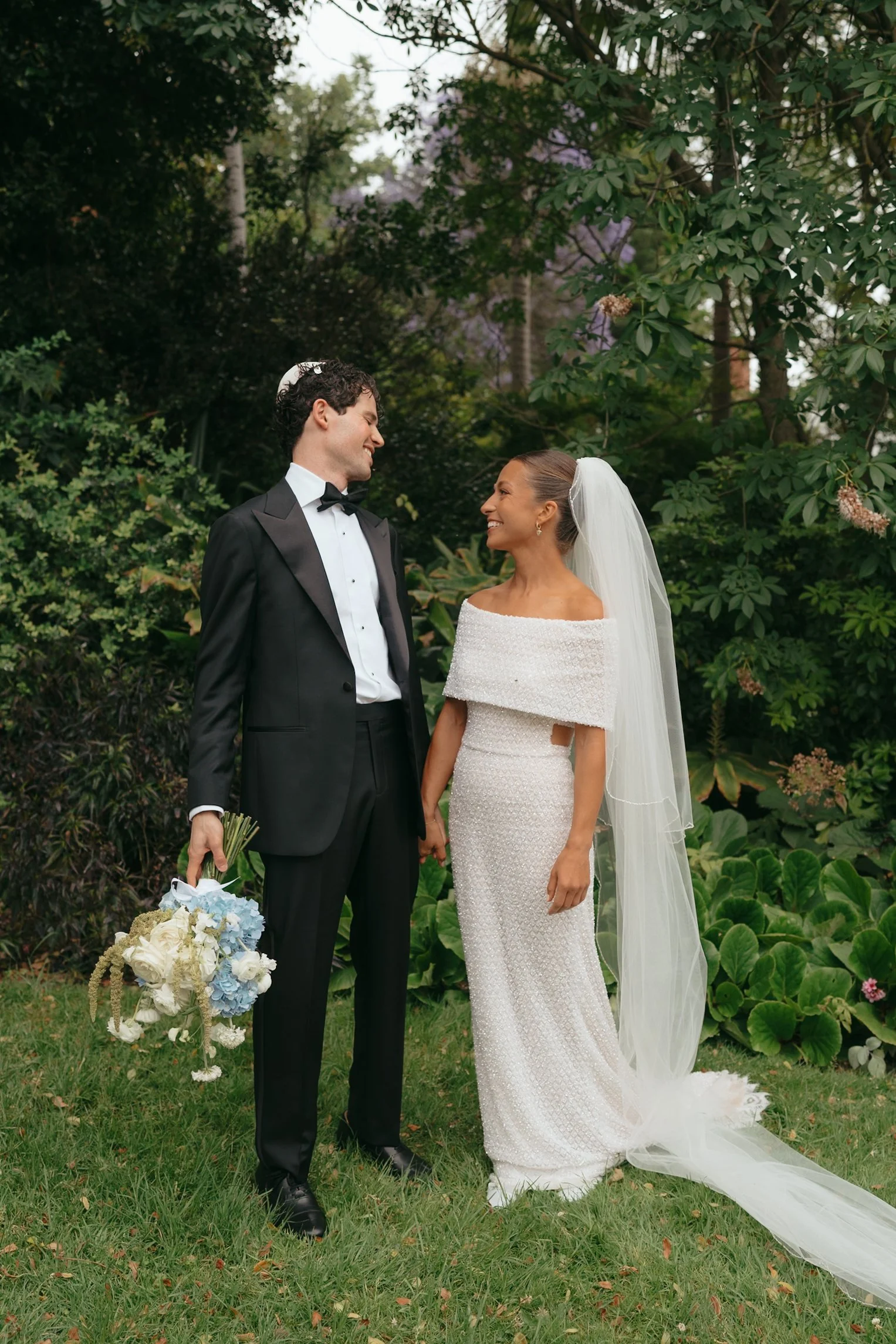A bride and groom standing together outdoors, smiling at each other in a garden setting, surrounded by greenery and trees.