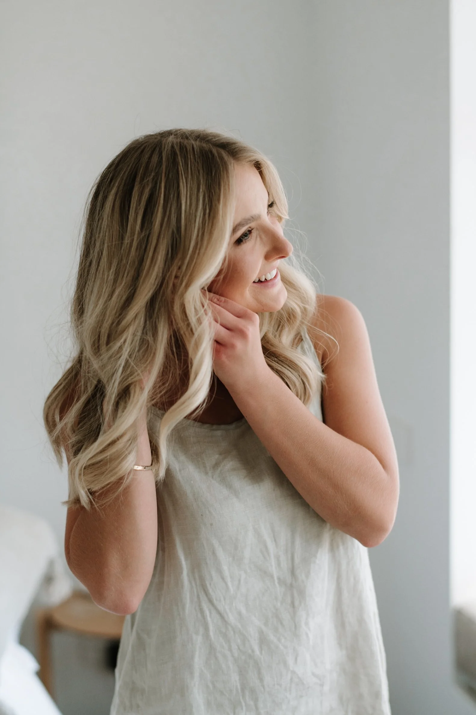 A young woman with long blonde hair smiling and looking to her right, wearing a sleeveless beige top, in a softly lit room with a white wall background.