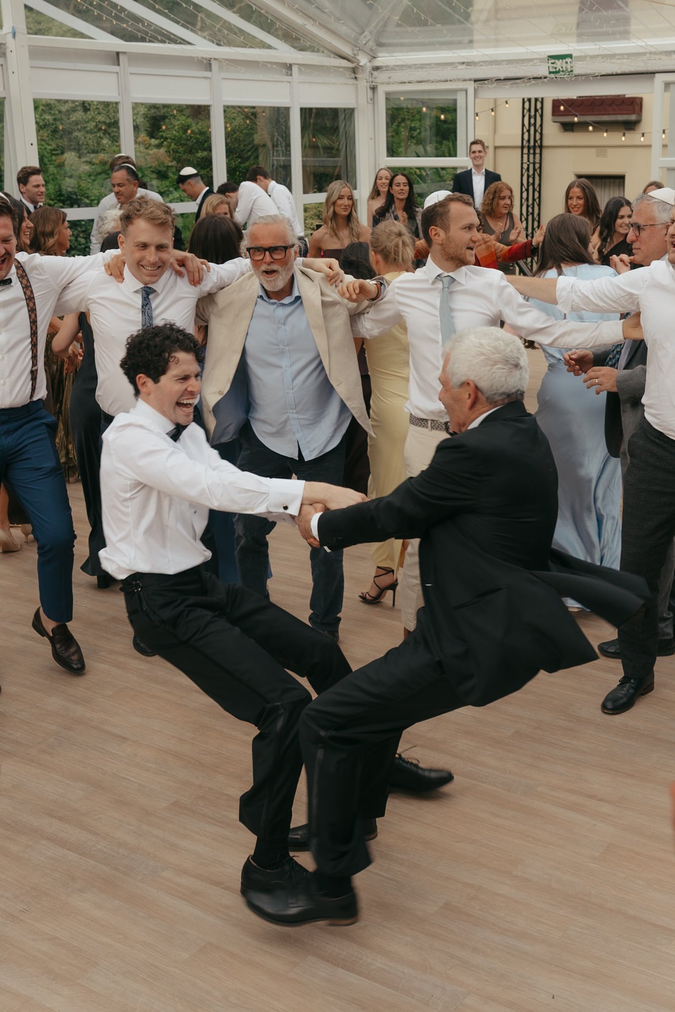 Group of people enjoying a lively dance at a celebration or party, some holding hands and others playing or dancing energetically.