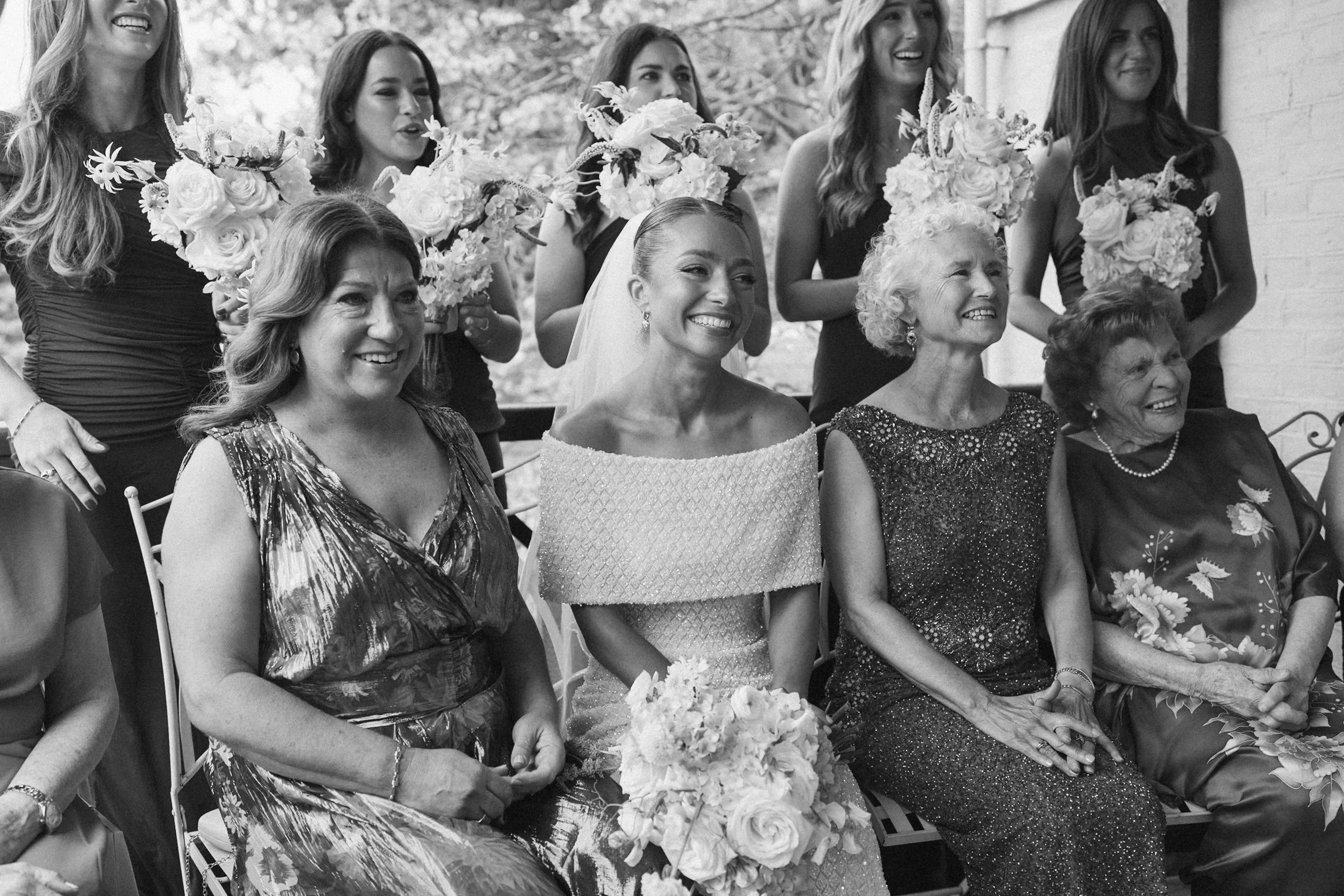 A group of women, including a bride with a veil, and bridesmaids holding bouquets, gathered for a wedding celebration, sitting outdoors on chairs.