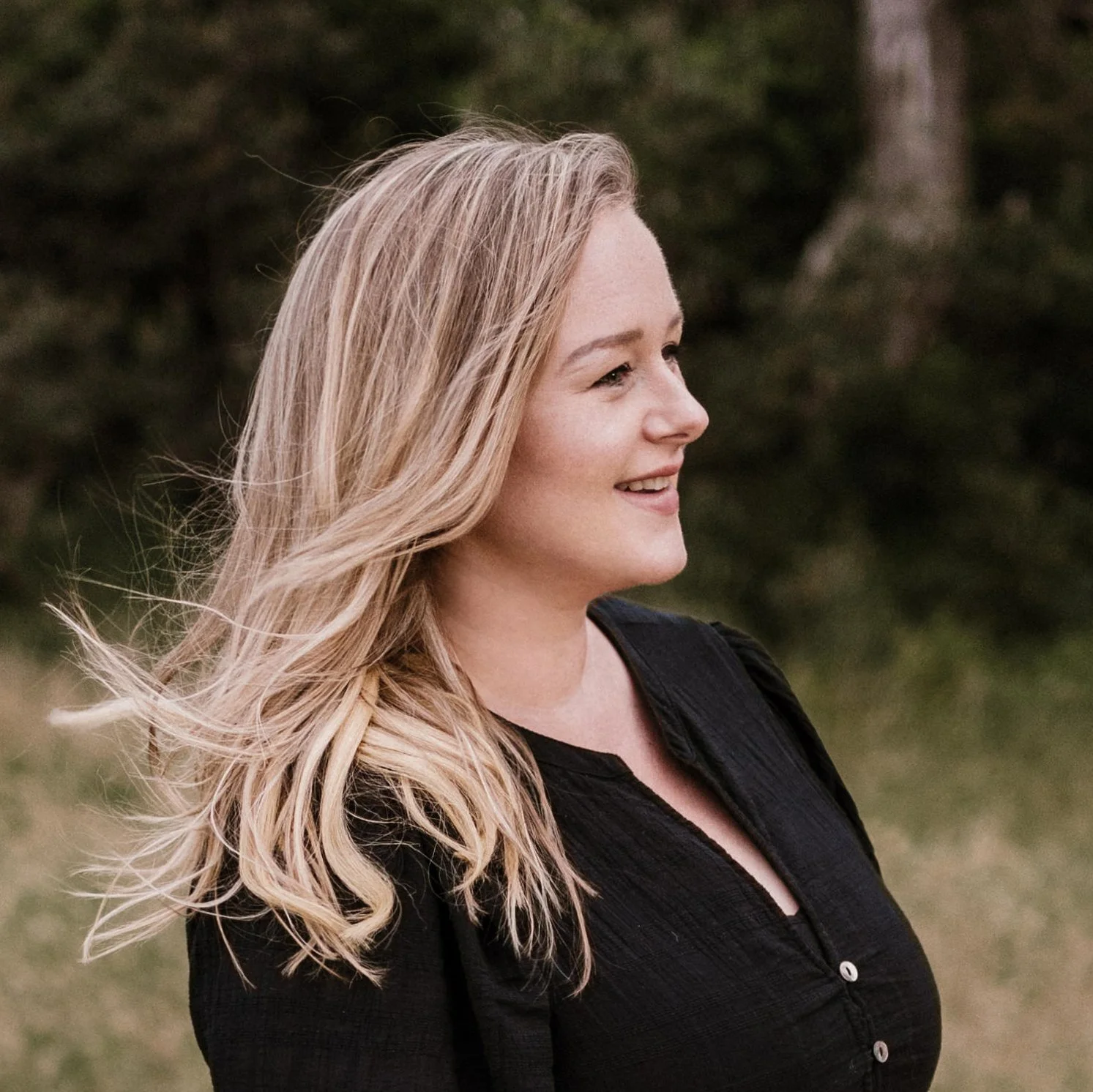 A young woman with long, wavy blonde hair is smiling and looking to the right outdoors, wearing a black top and standing against a natural background with trees and grass.