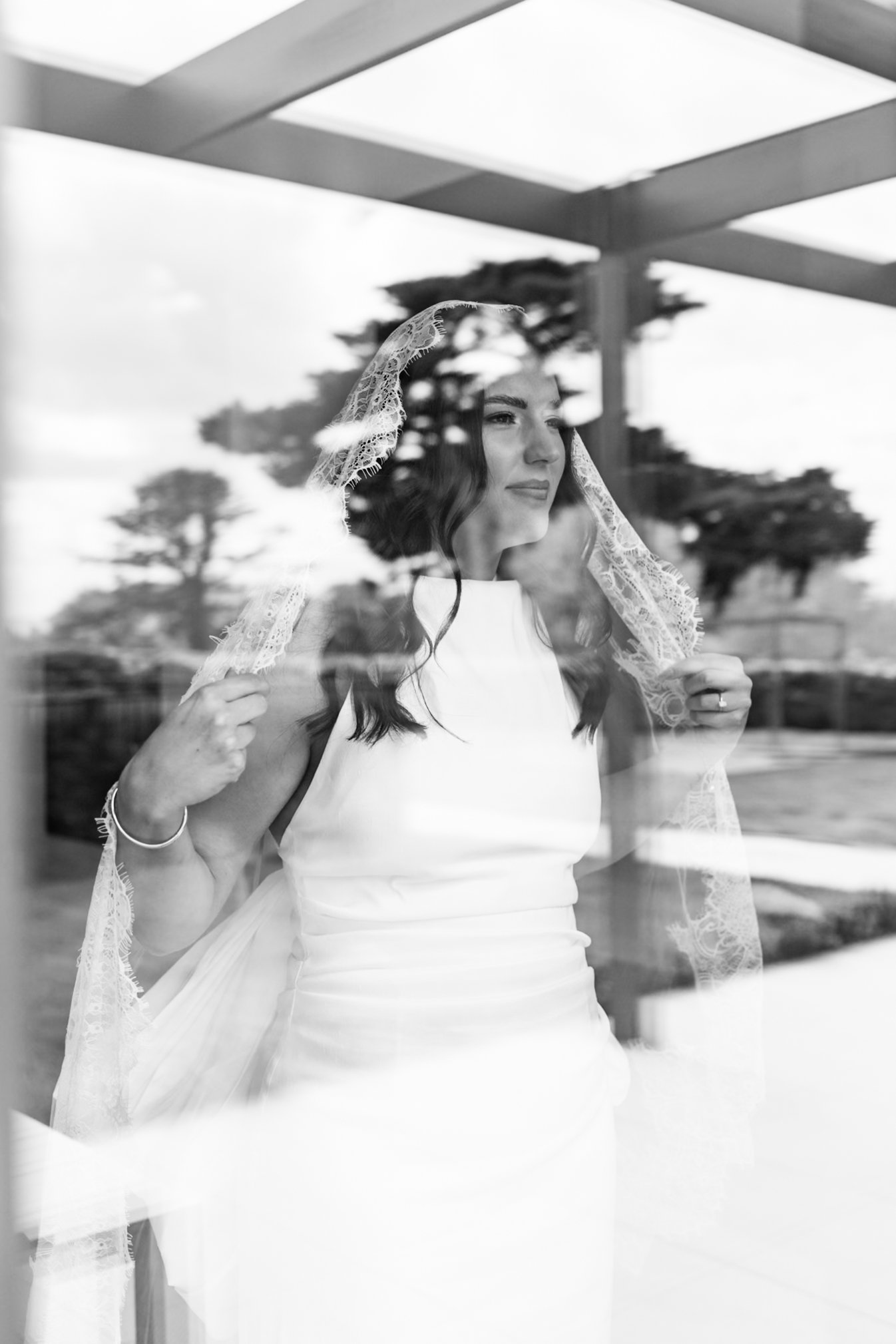 A woman in a wedding dress with lace veil looking out a window, reflection visible, trees and sky outside.