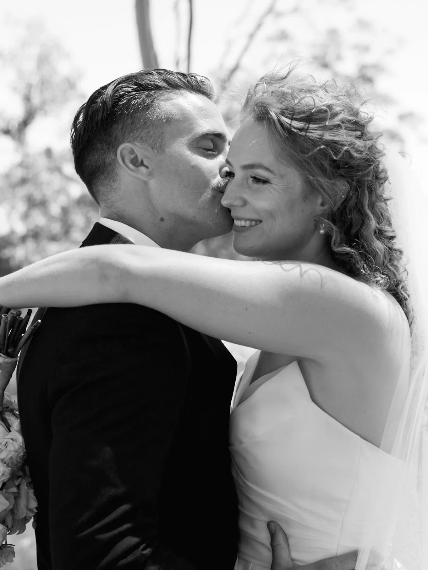 A black and white photograph of a bride and groom embrace and share a kiss during their wedding, with the groom in a dark suit and the bride in a white wedding dress.