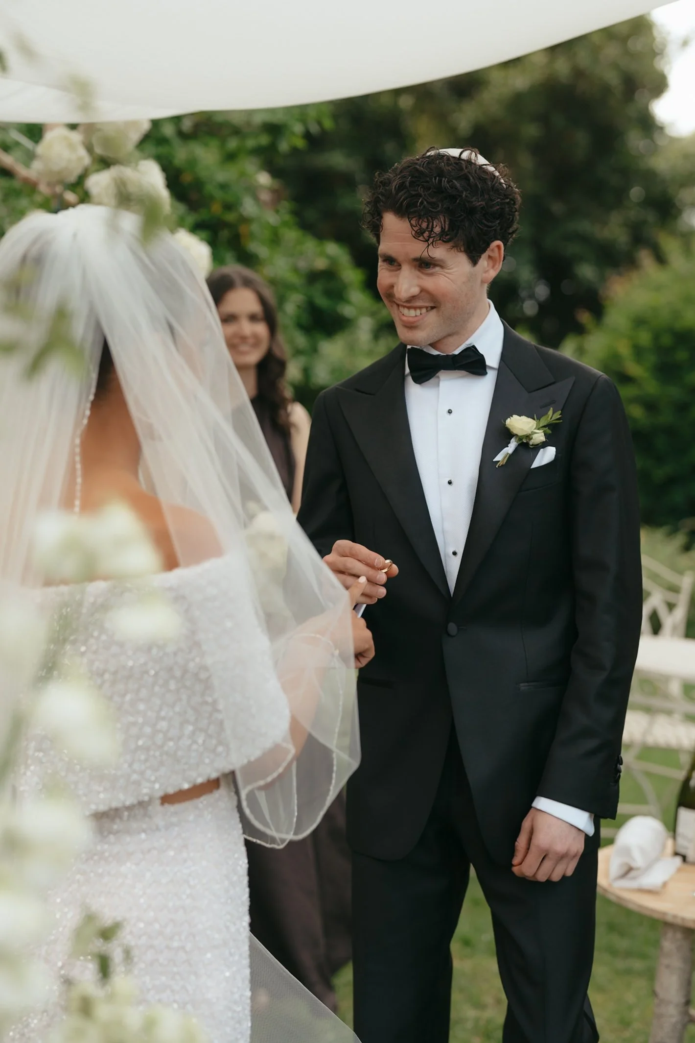 A groom in a black tuxedo and bow tie smiling at a bride during their outdoor wedding ceremony. The bride is wearing a white dress with beads and a veil. A woman watching is visible in the background, and there are green trees and floral decorations 